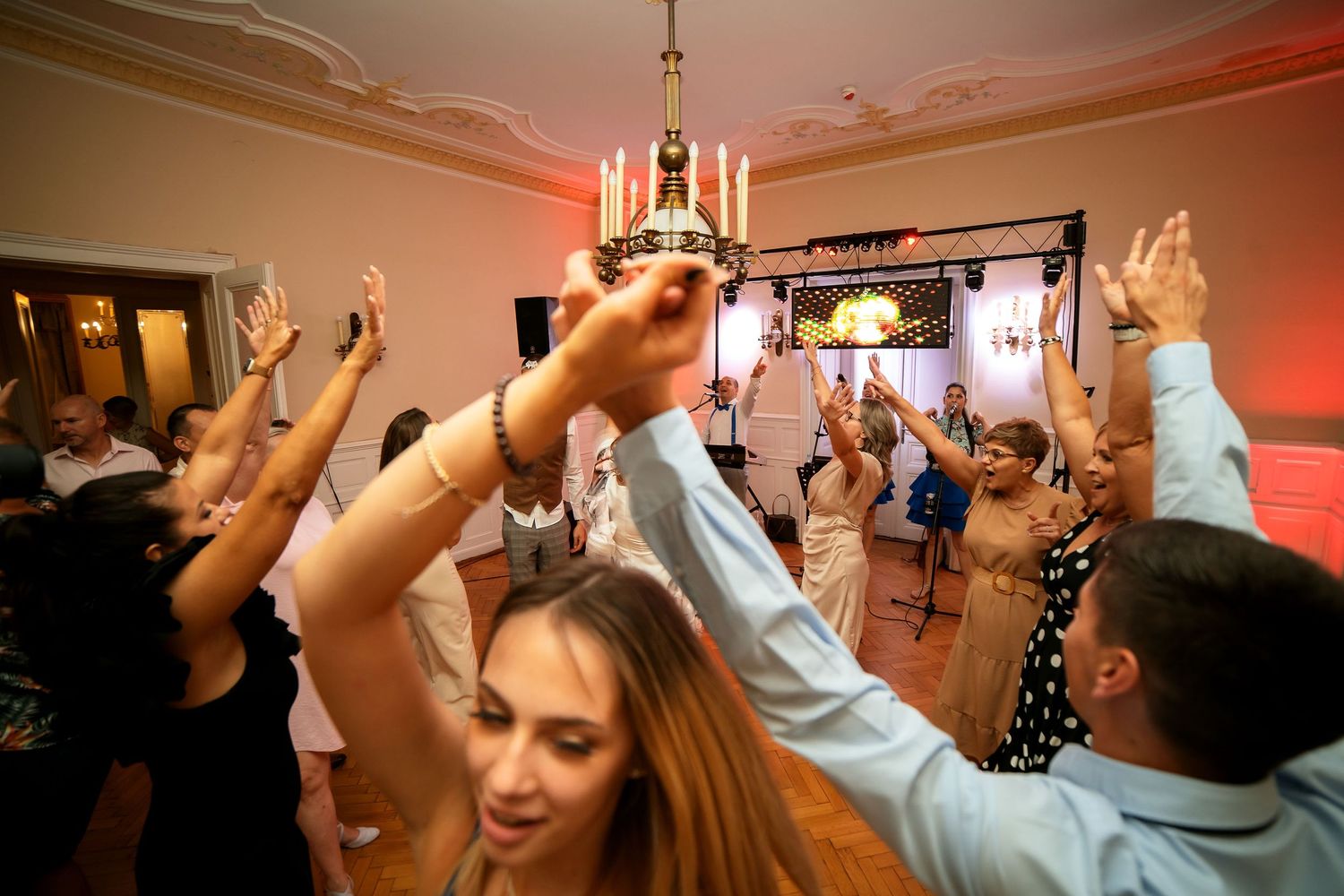 Party guests dancing with raised arms under an elegant chandelier at an indoor celebration event.