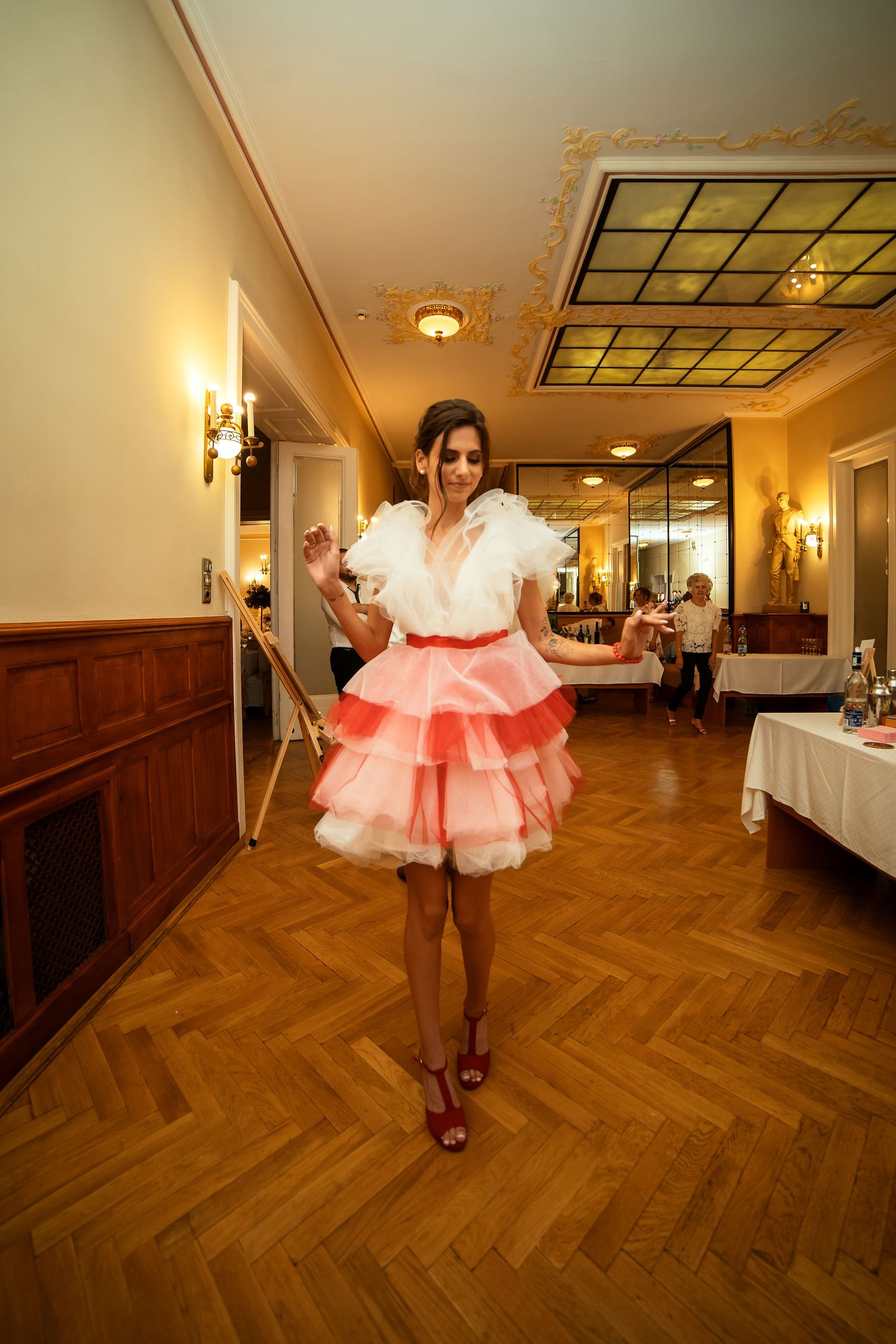Person wearing a white ruffled top and pink layered skirt poses in an elegant venue hallway.