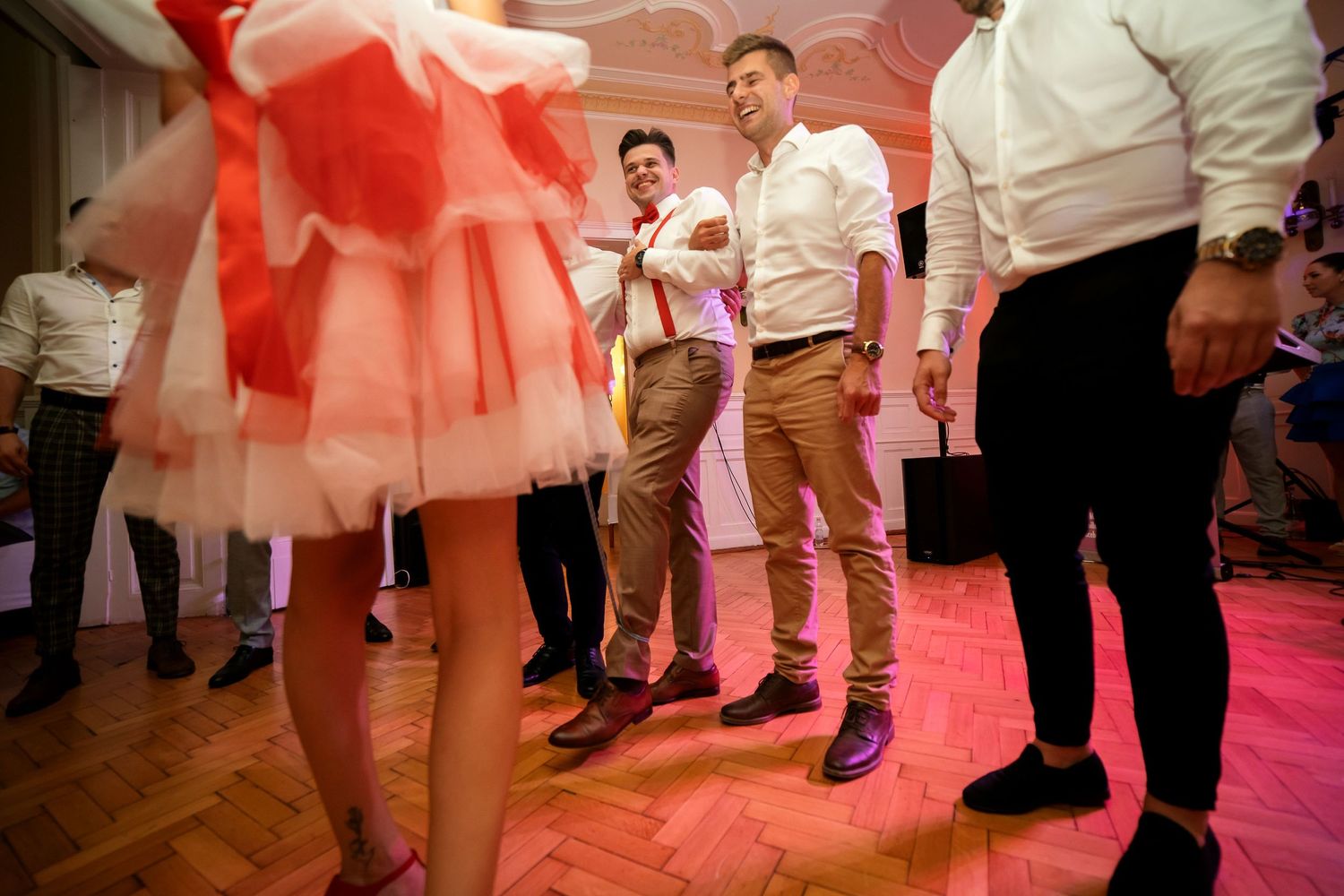 Group of party guests dancing on a parquet floor under pink lighting.