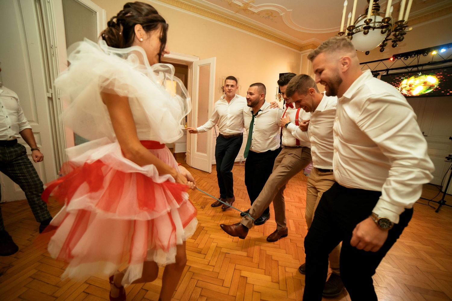 Energetic dancers in white shirts gather on a parquet floor around someone wearing a red and white ruffled dress.
