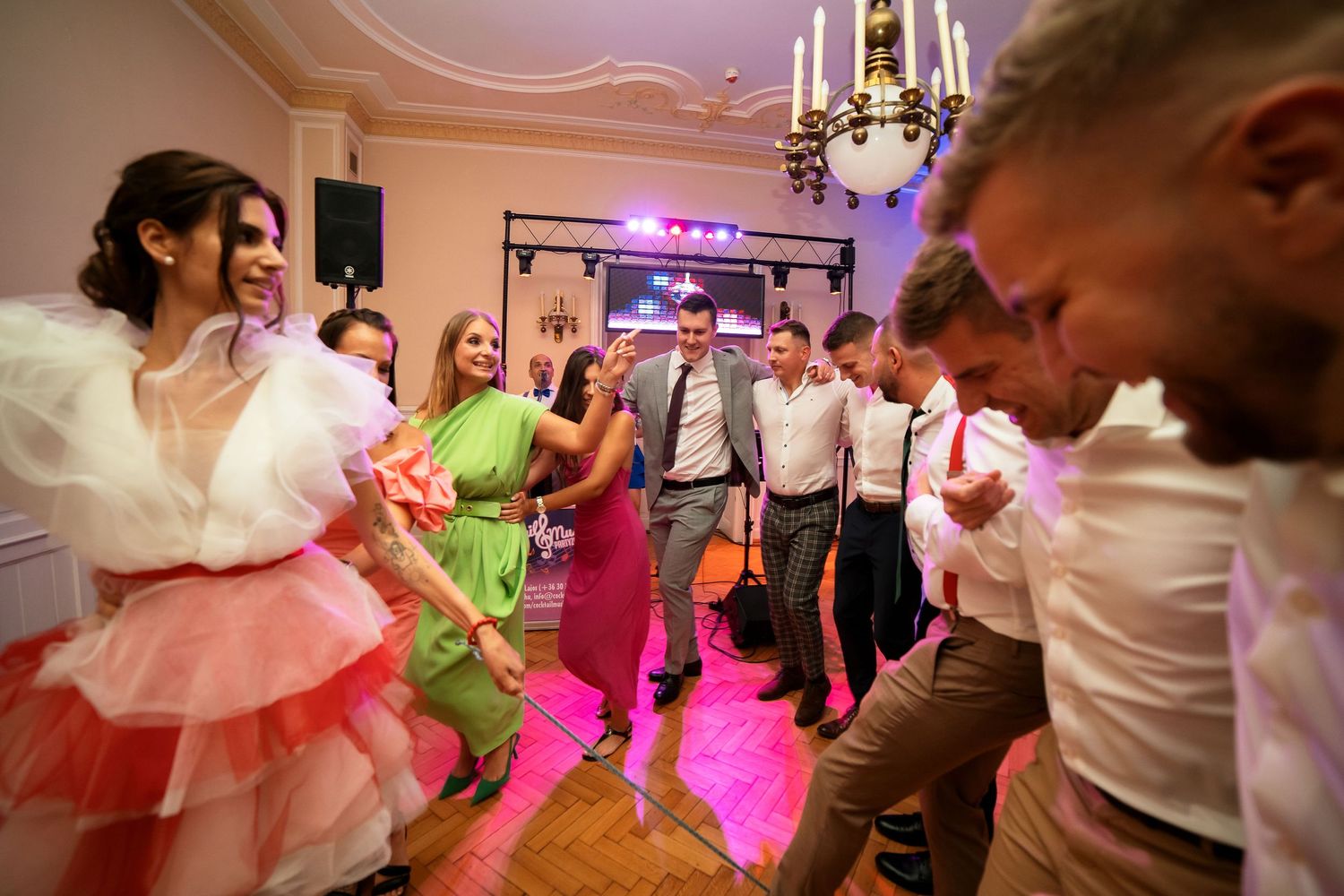 Colorful party lights illuminate dancers on a wooden floor with chandelier overhead.
