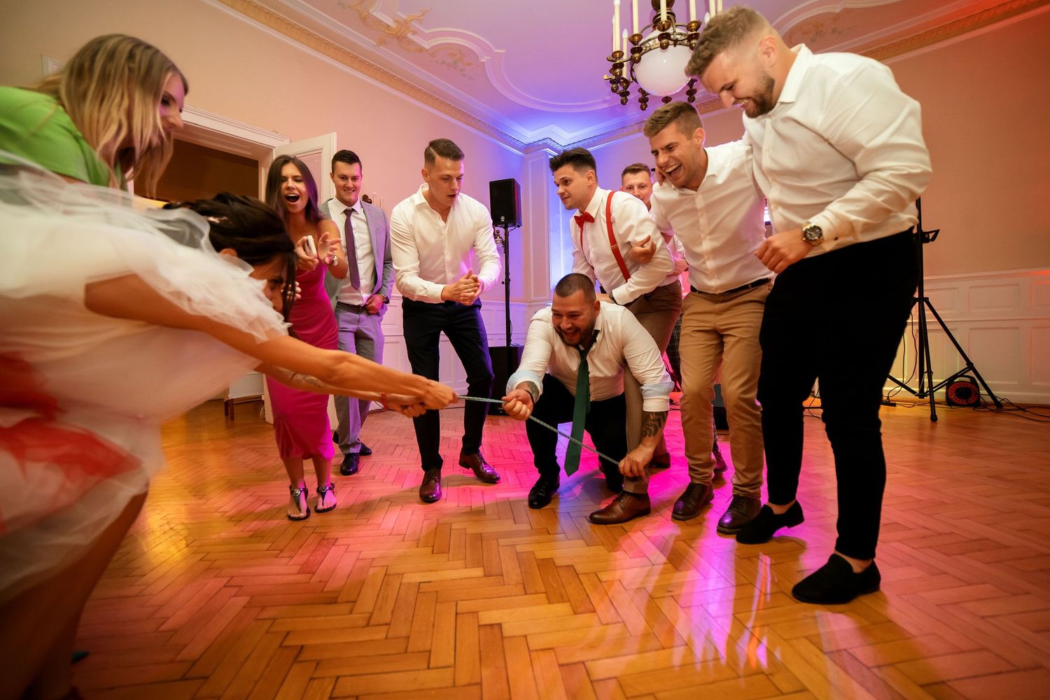 Dance party participants perform synchronized moves on a wooden floor under pink lighting.