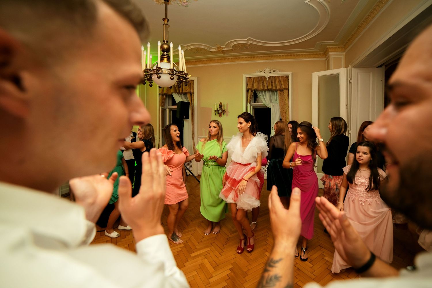 Energetic wedding guests dance together in colorful dresses at an elegant indoor reception venue.