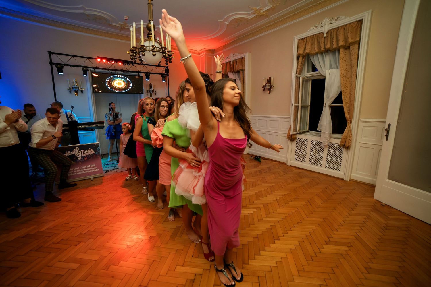 Wedding party dances under dramatic red and blue lighting in an elegant ballroom with parquet floors.