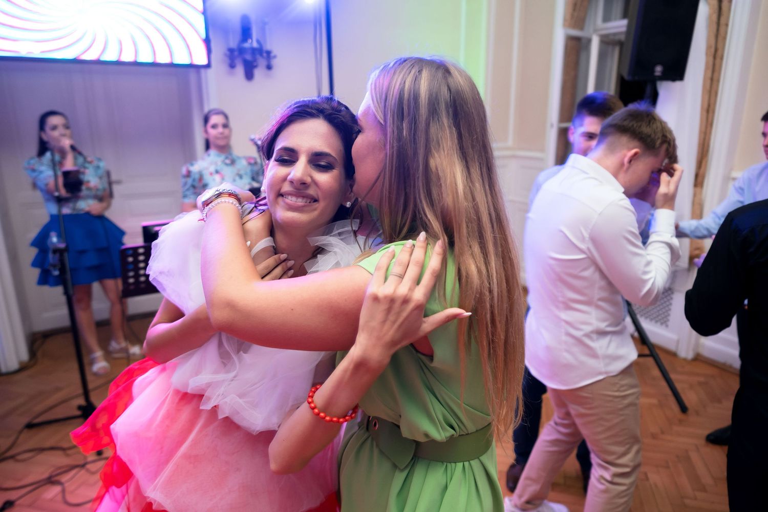 Two people dancing together energetically at a party in colorful dresses with other guests visible in background.