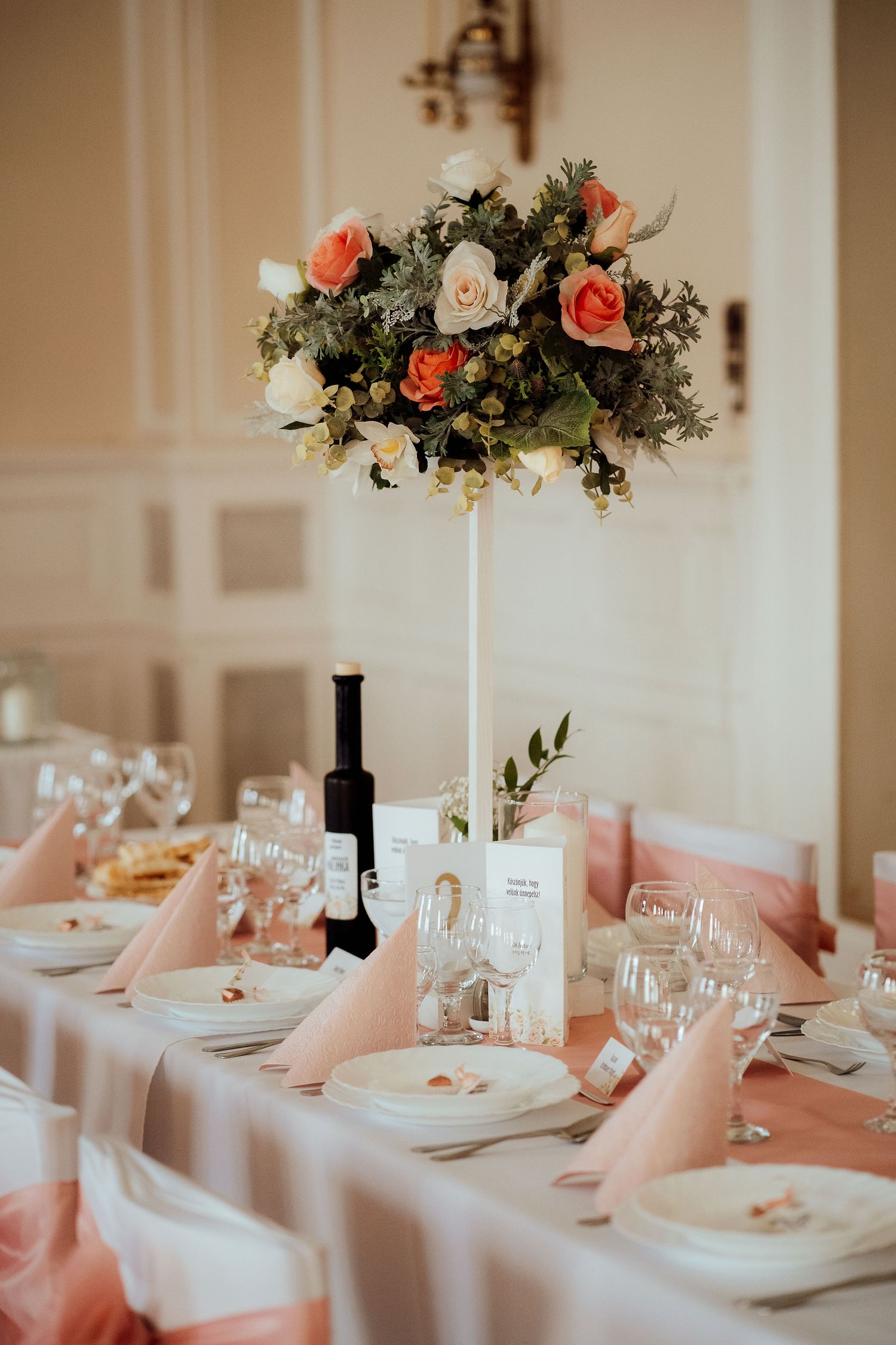 Formal table setting with tall floral arrangement, wine bottle and peach accents.