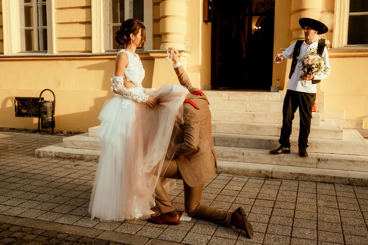 A couple performs a playful wedding photo pose with theatrical flair outside a yellow building.