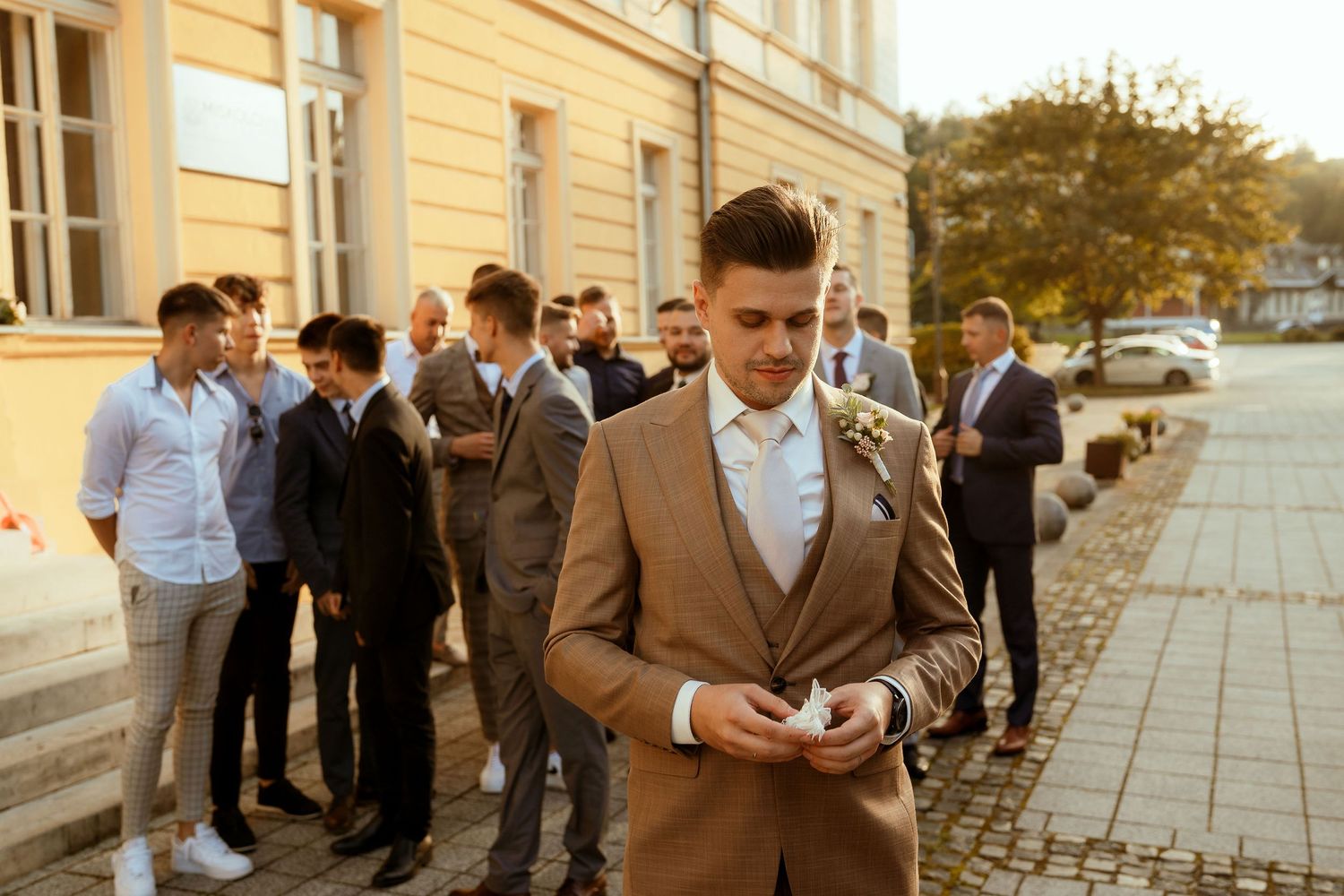 A stylishly dressed guest in a brown suit stands outside during a wedding event.
