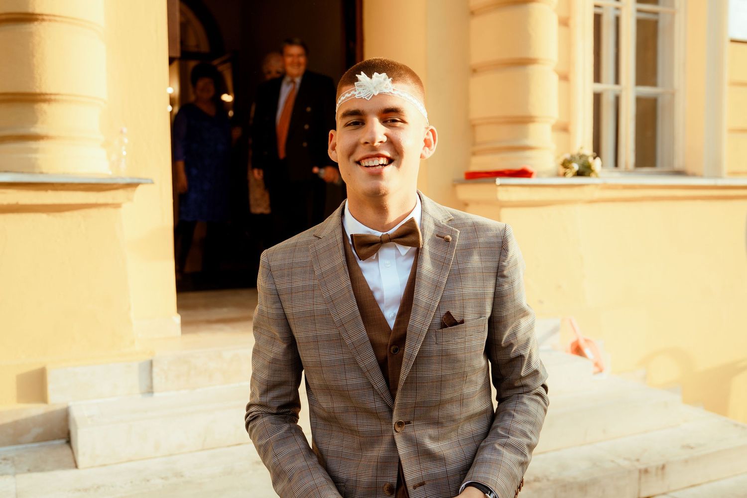 A guest in a grey suit and bowtie smiles warmly outside a yellow building.