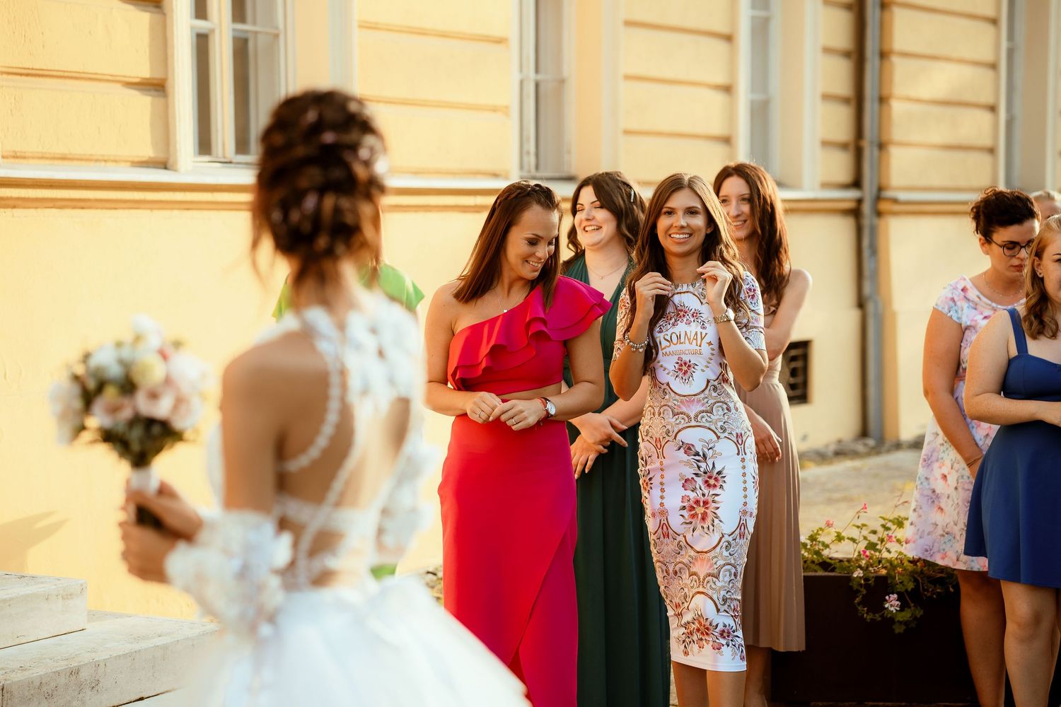 Wedding guests in colorful dresses gather to greet the bride outside.