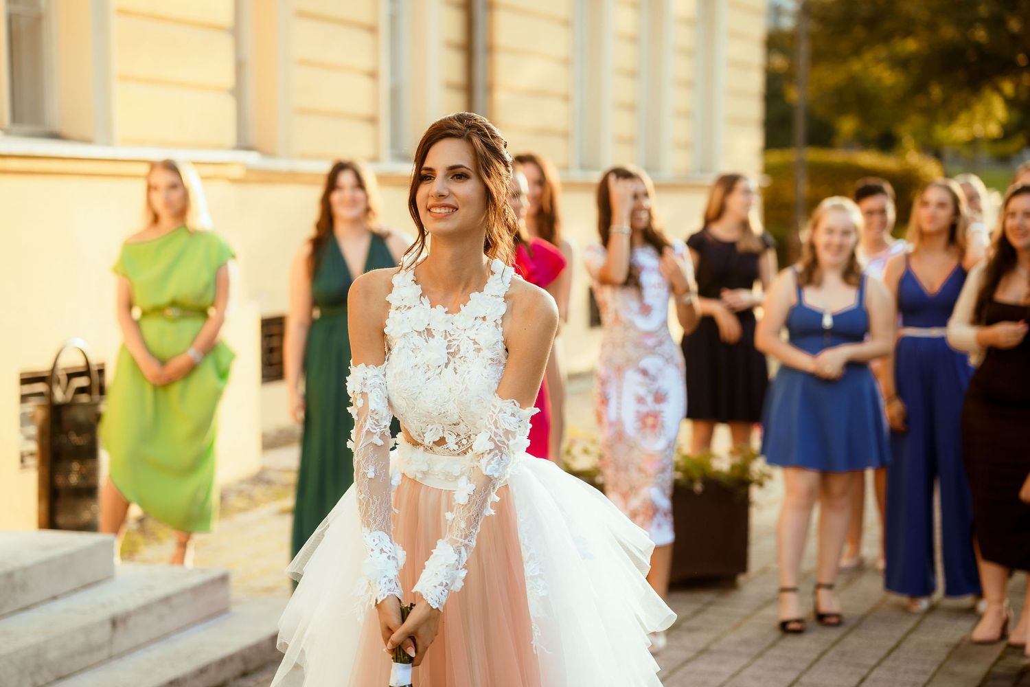 A bride in a delicate lace wedding dress stands before guests on stone steps.