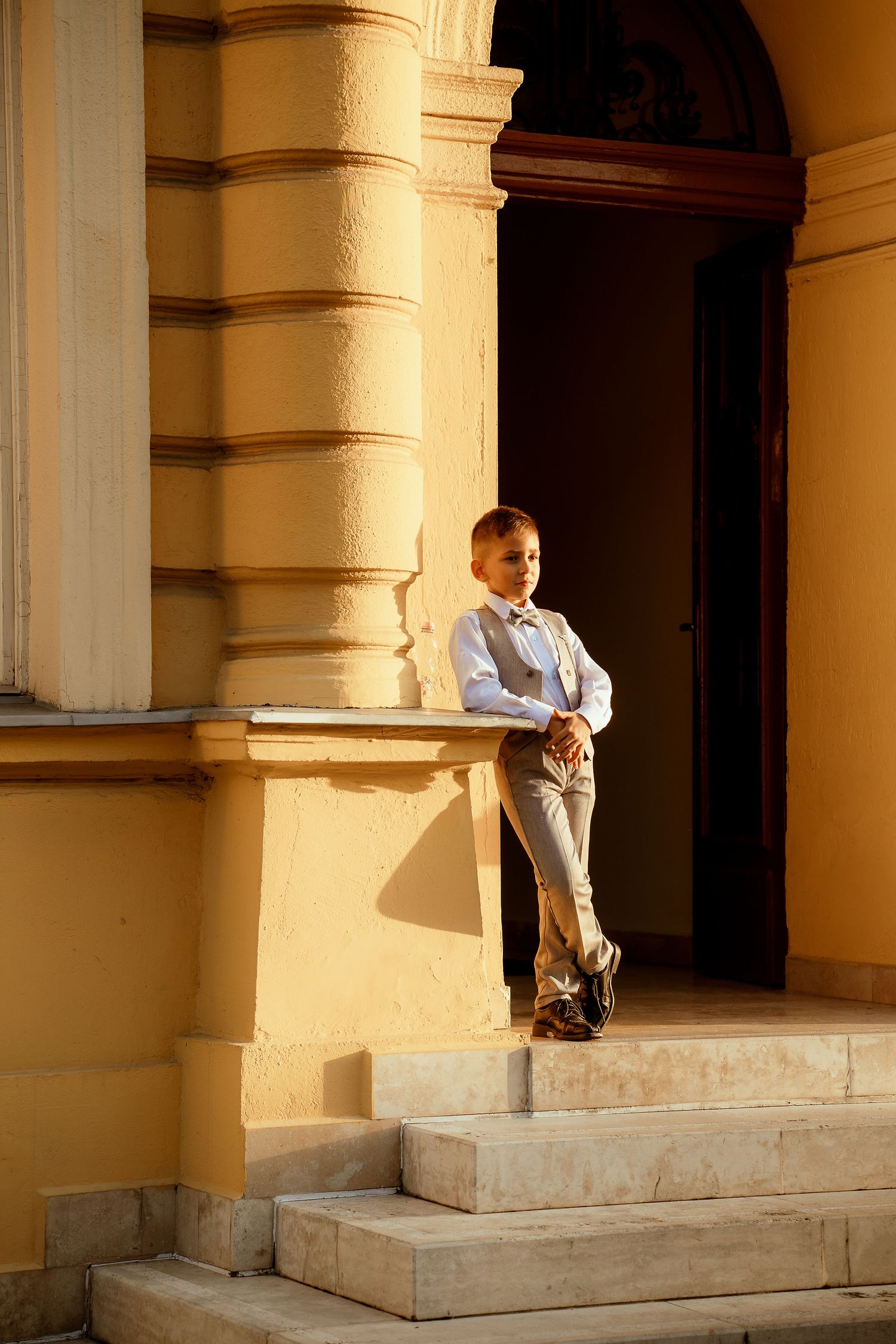 A young attendee in formal attire poses on stone steps of a yellow building.
