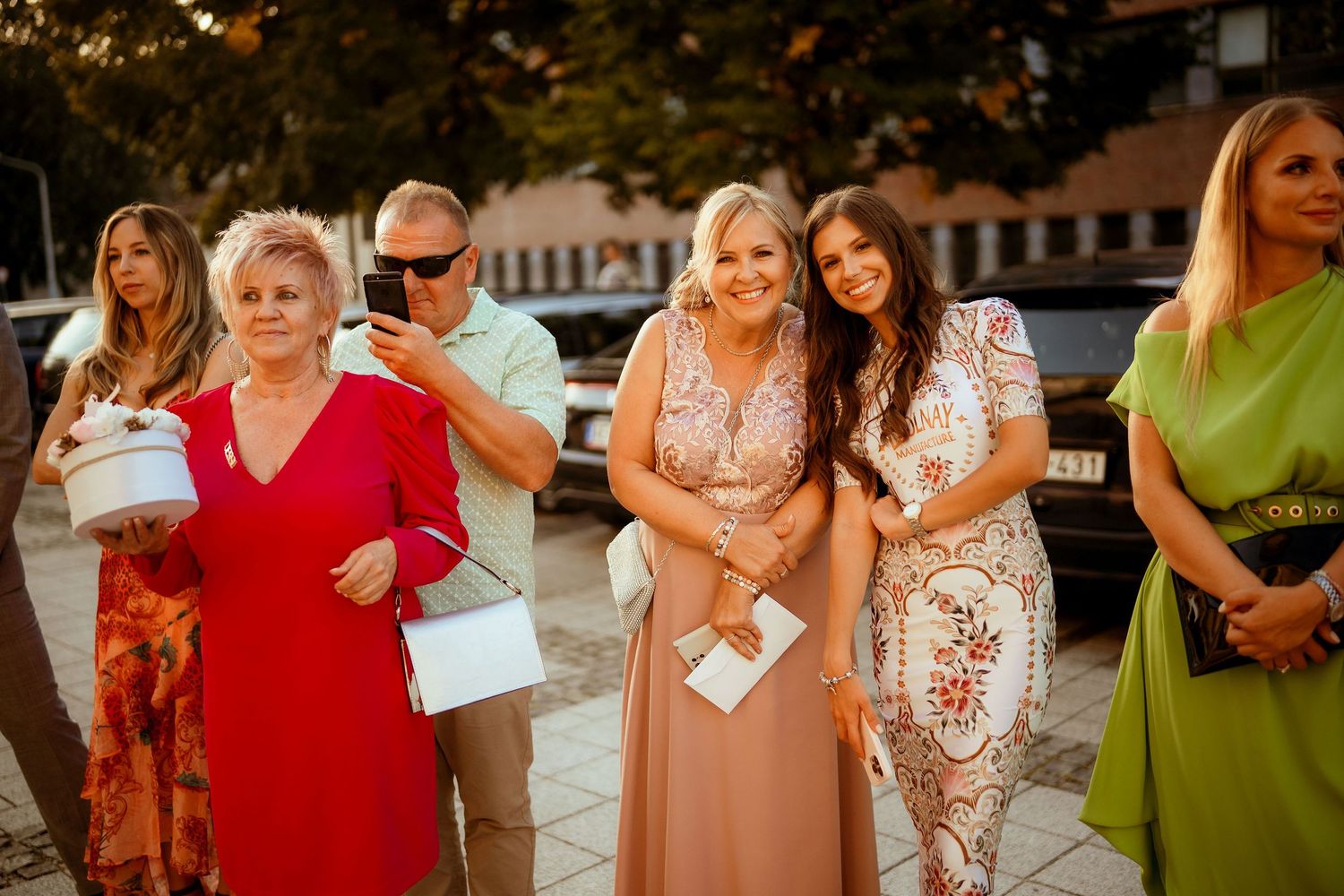 Wedding guests in colorful formal dresses gather outside during golden hour at an outdoor celebration.