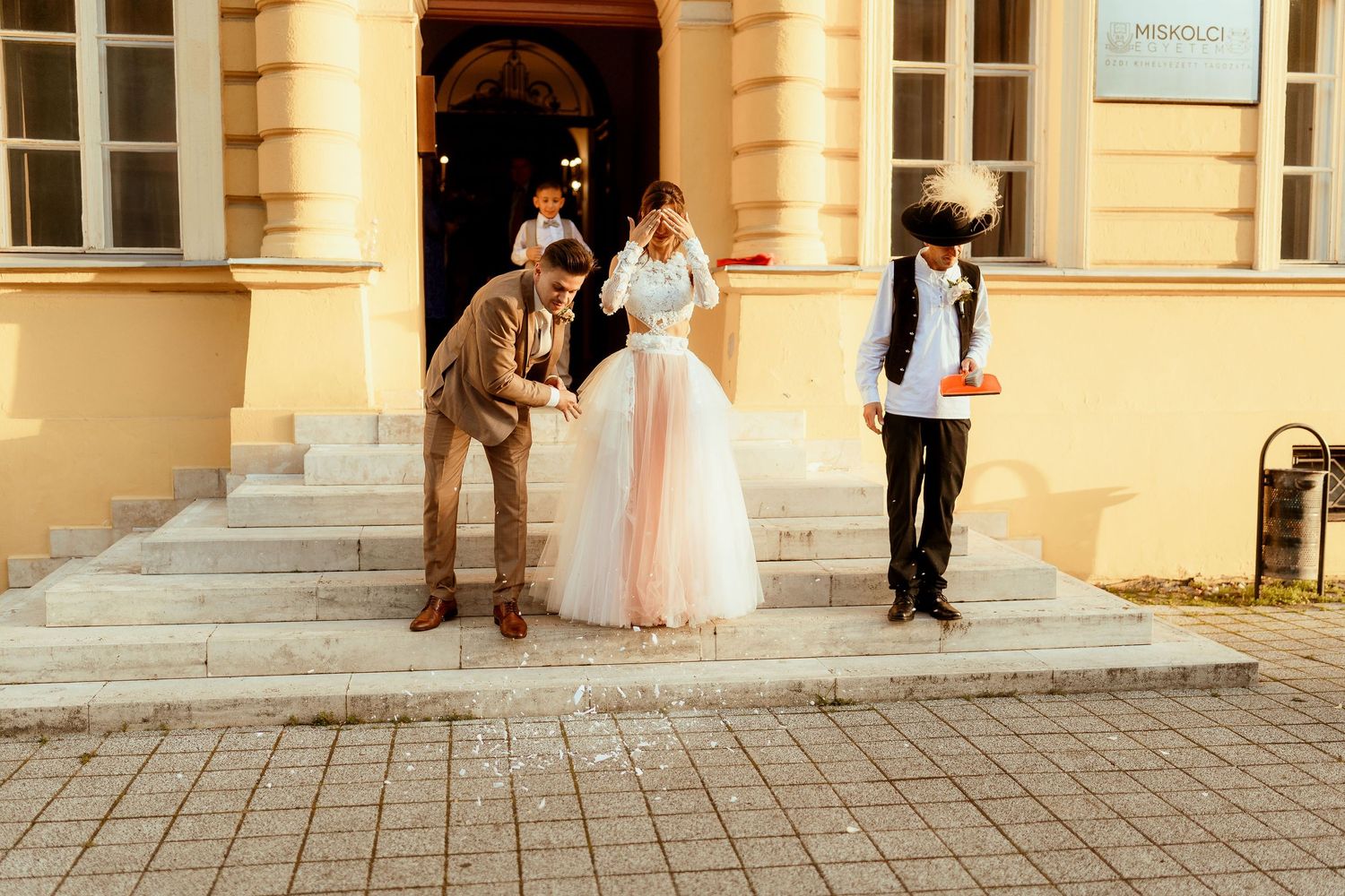A bride in a light pink tulle gown poses on historic building steps with guests.