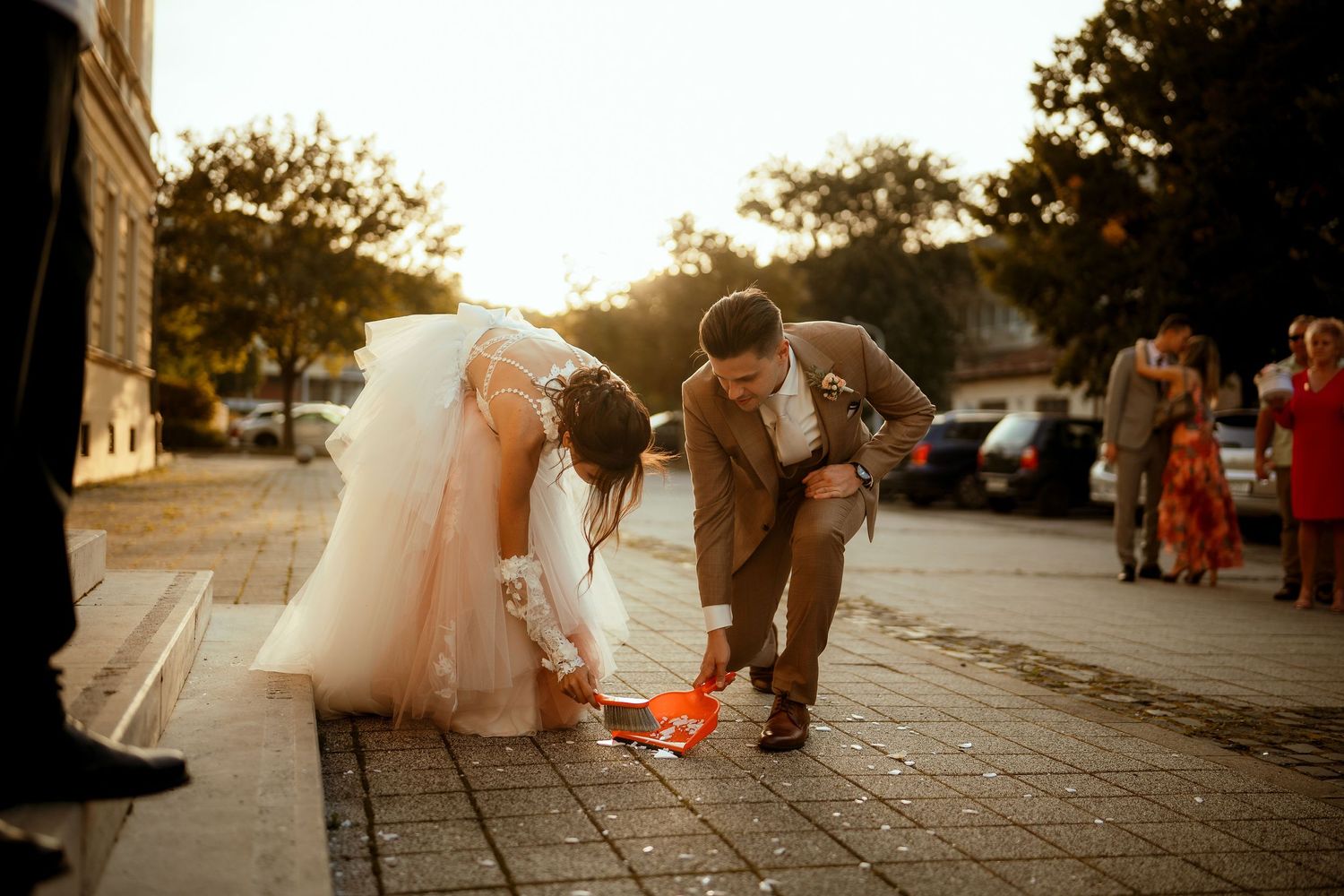 A couple bends down to perform a wedding tradition on cobblestone pavement at sunset.