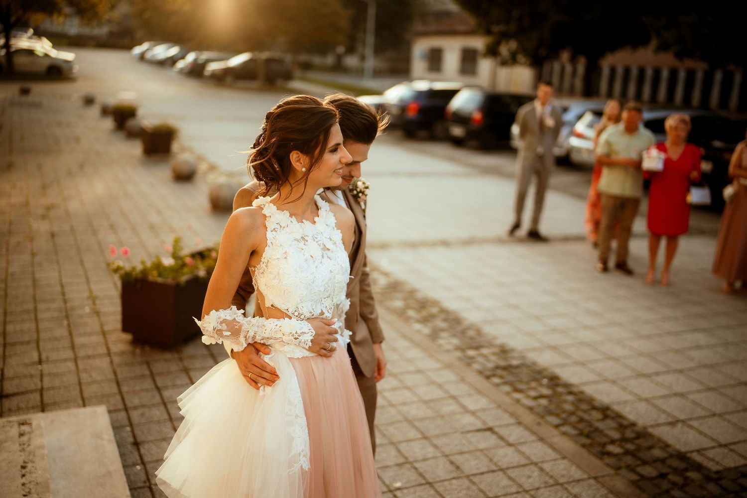 A sunny outdoor wedding celebration with guests in formal attire on a brick-paved street.