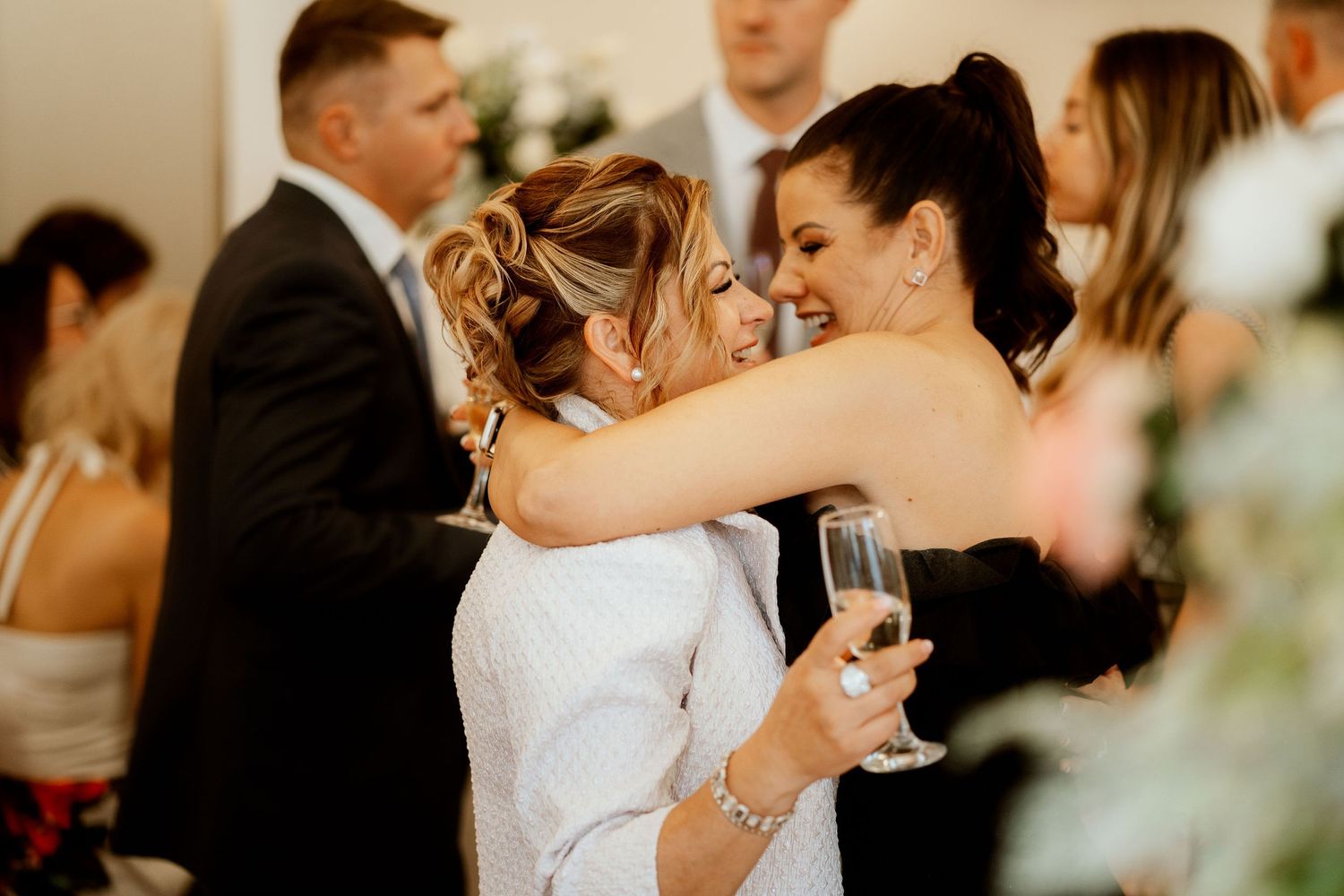 Wedding guests sharing embraces while holding champagne glasses at an indoor celebration.