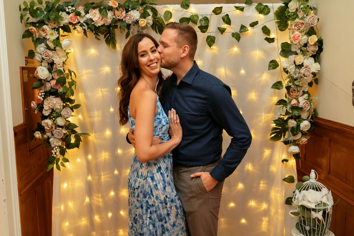 Couple poses under a romantic floral arch with twinkling lights and roses.