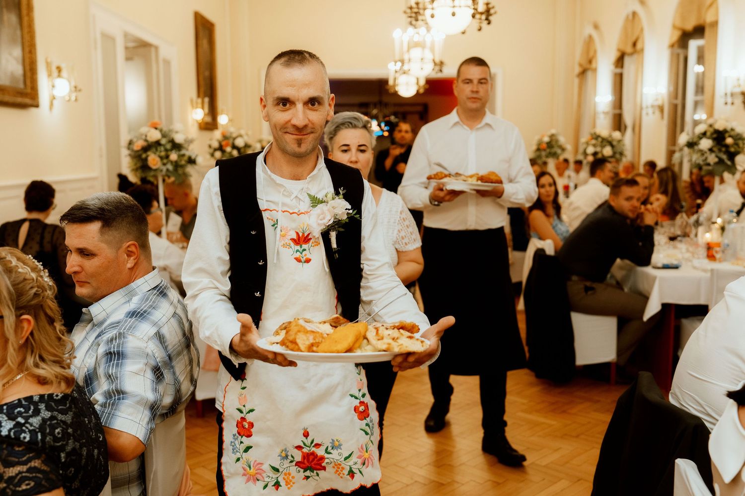 Server wearing floral apron carries plates through elegant reception hall.