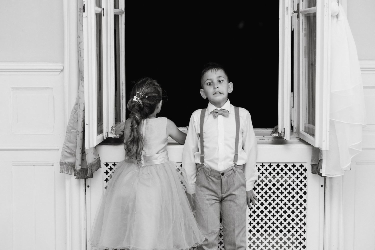 Two children in formal attire stand at a doorway in black and white.