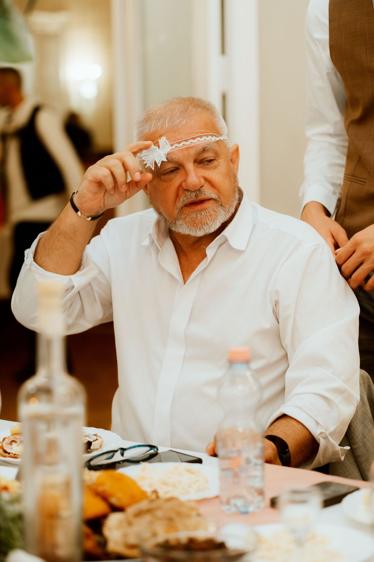 A guest at a wedding reception wipes his forehead while seated at a dining table.