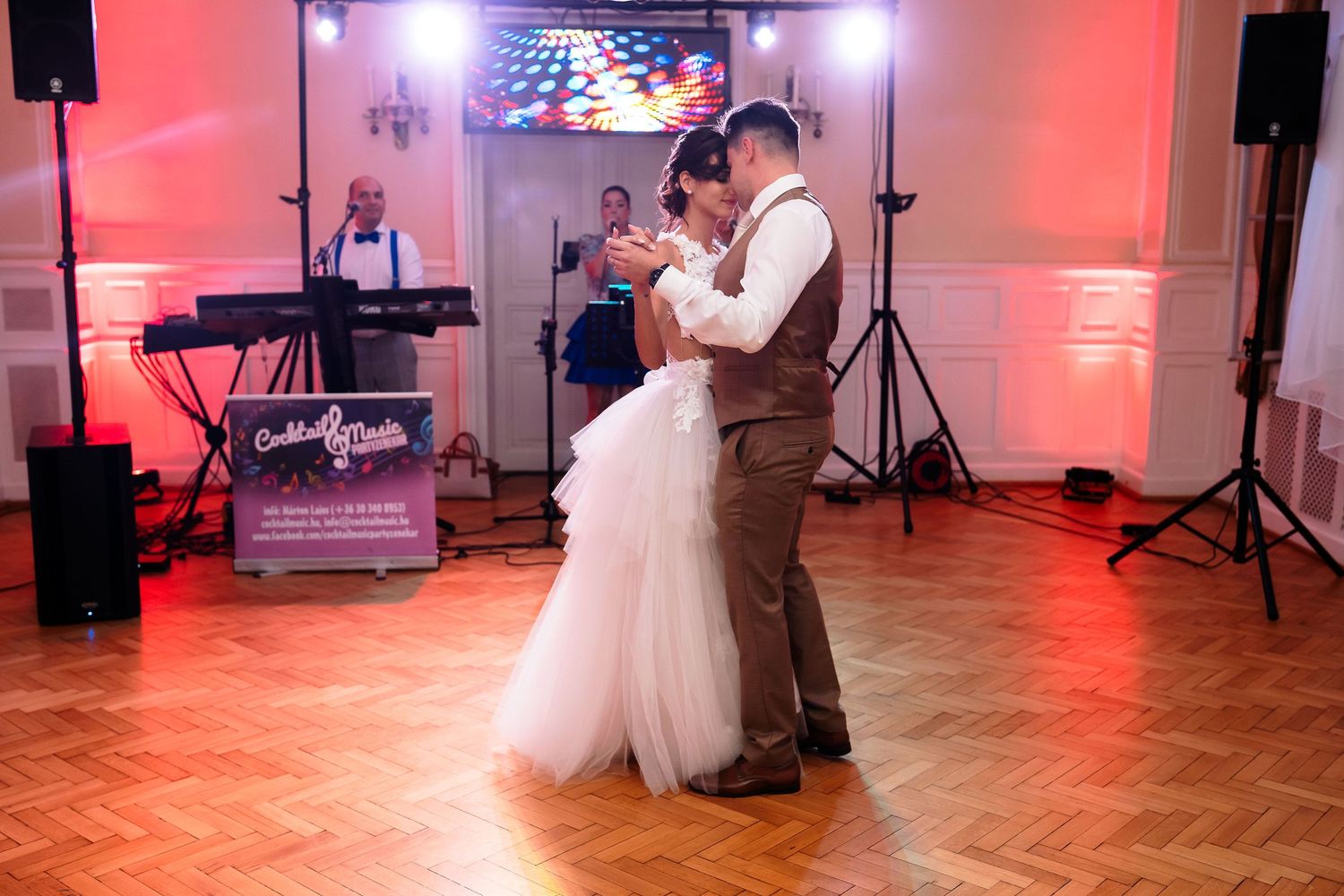 A couple shares their first dance on a wooden floor with pink ambient lighting and DJ equipment.