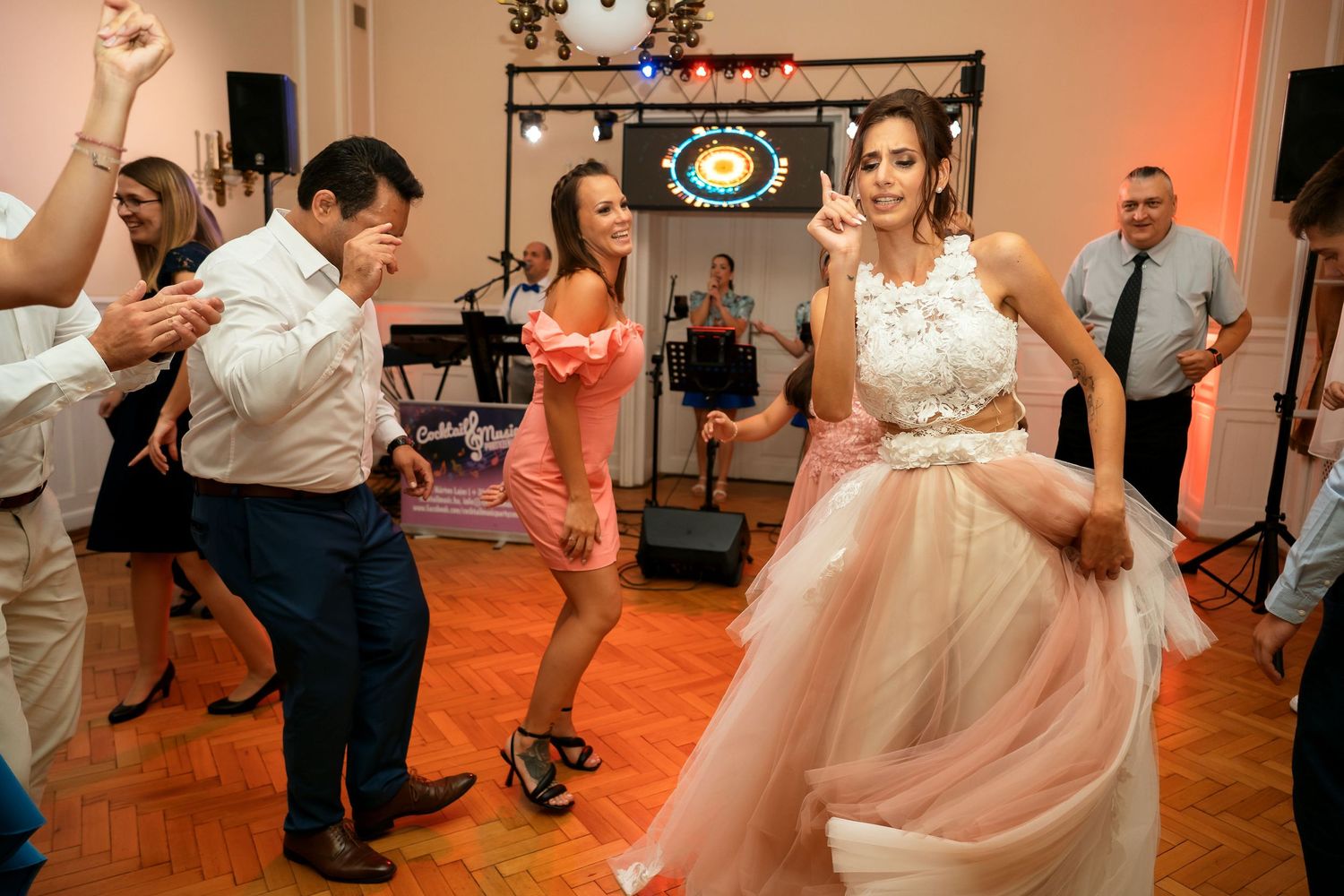 Wedding party dancing energetically on a wooden dance floor with ambient lighting.