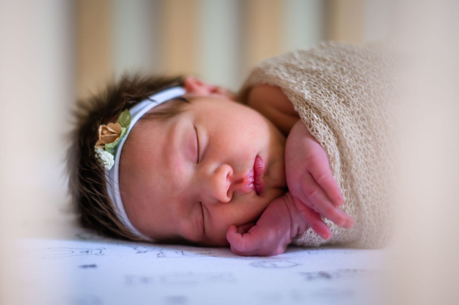 A peaceful sleeping newborn wearing a delicate headband wrapped in beige knit fabric.