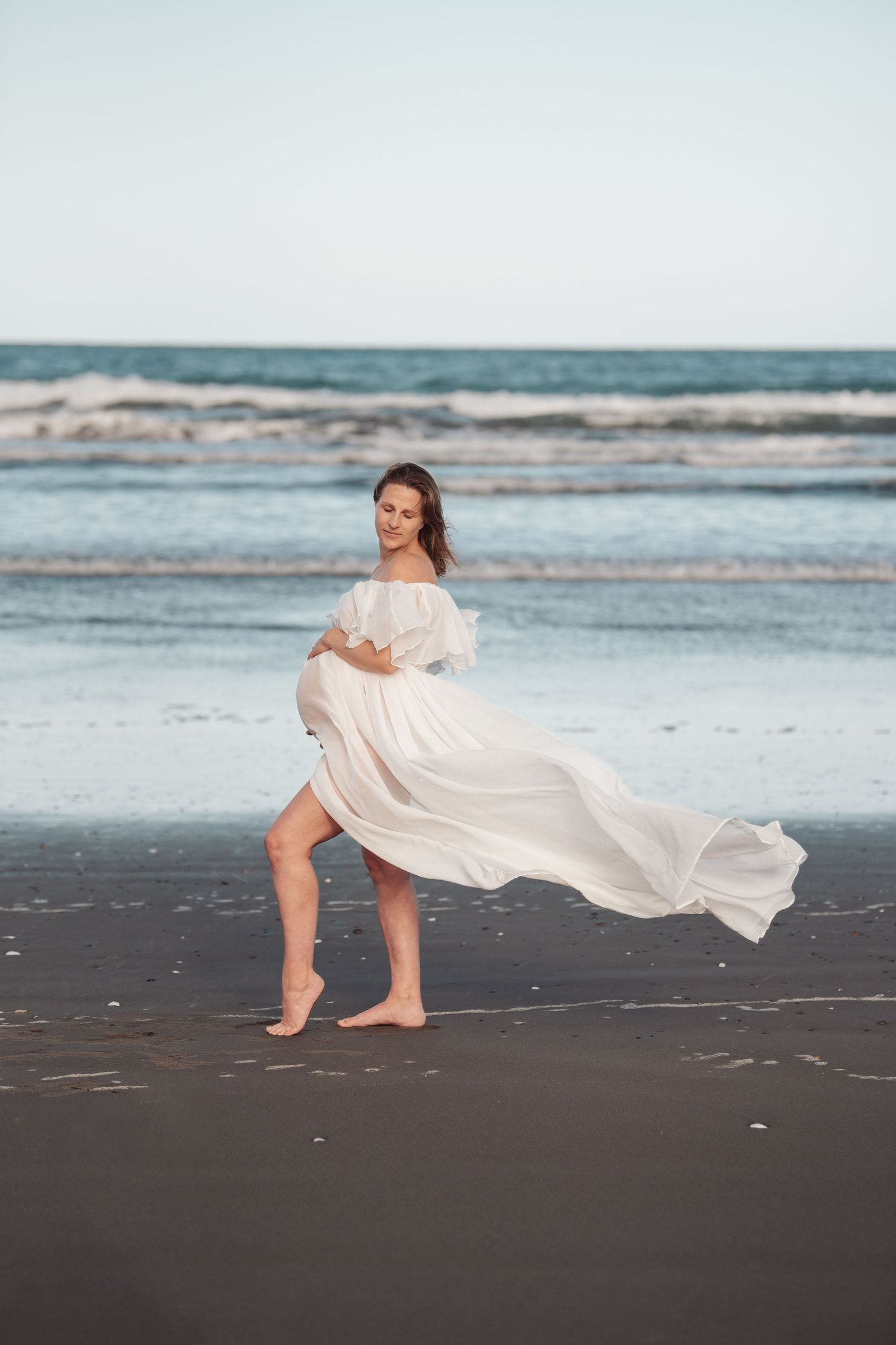 Flowing white dress billows in ocean breeze as woman walks barefoot on beach at sunset.