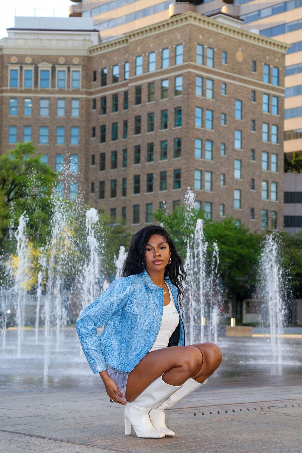 Woman in denim jacket crouches by fountain with modern architecture in background.