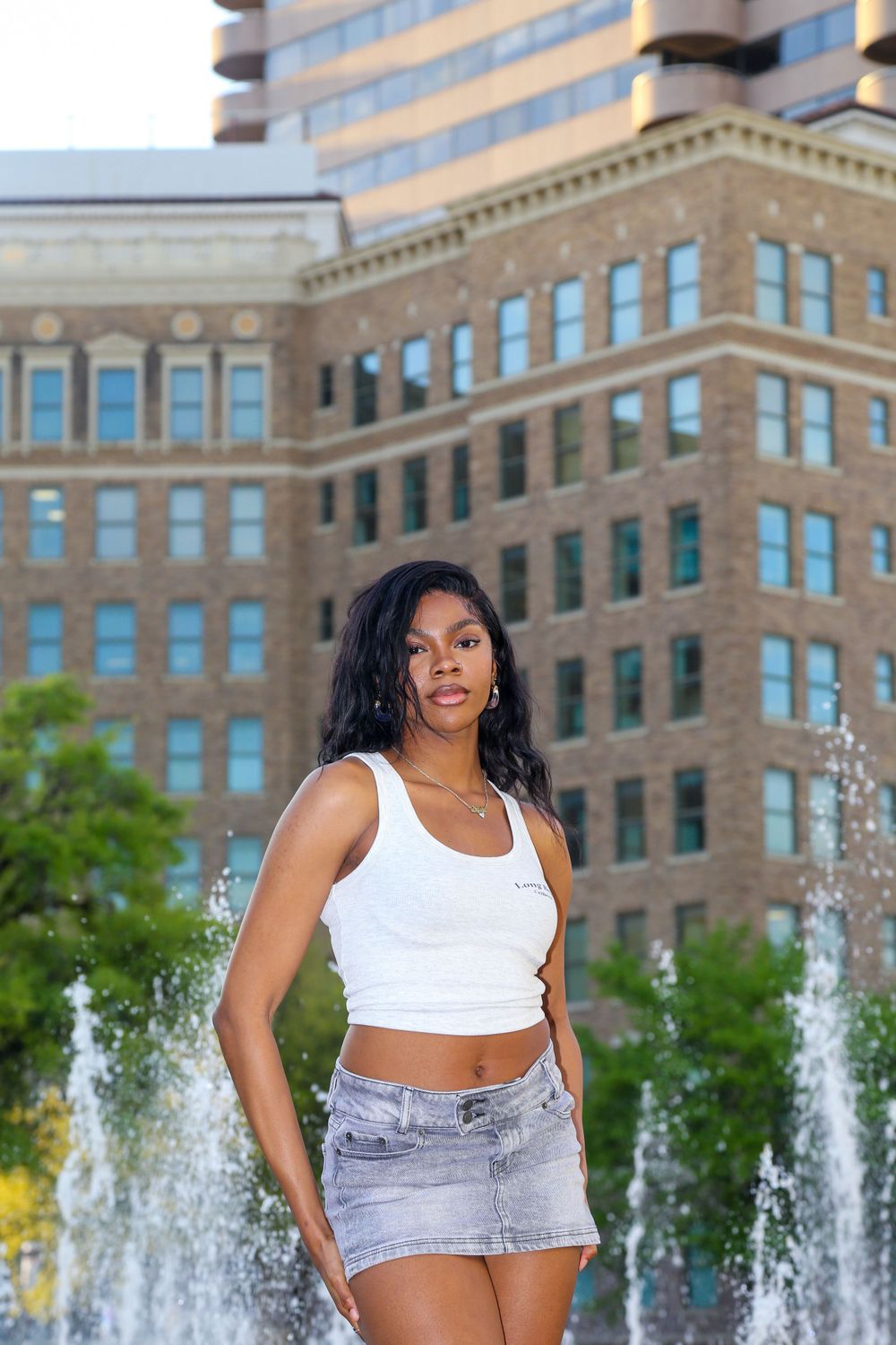 Woman in white crop top and denim shorts poses in front of urban fountain and building.