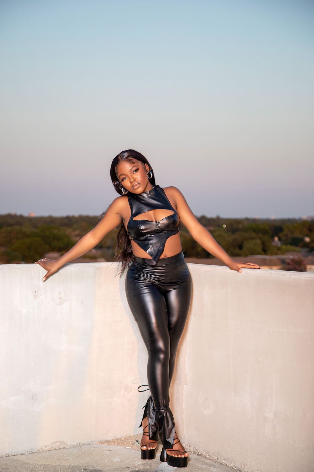 Woman in black leather outfit poses on white wall during sunset.