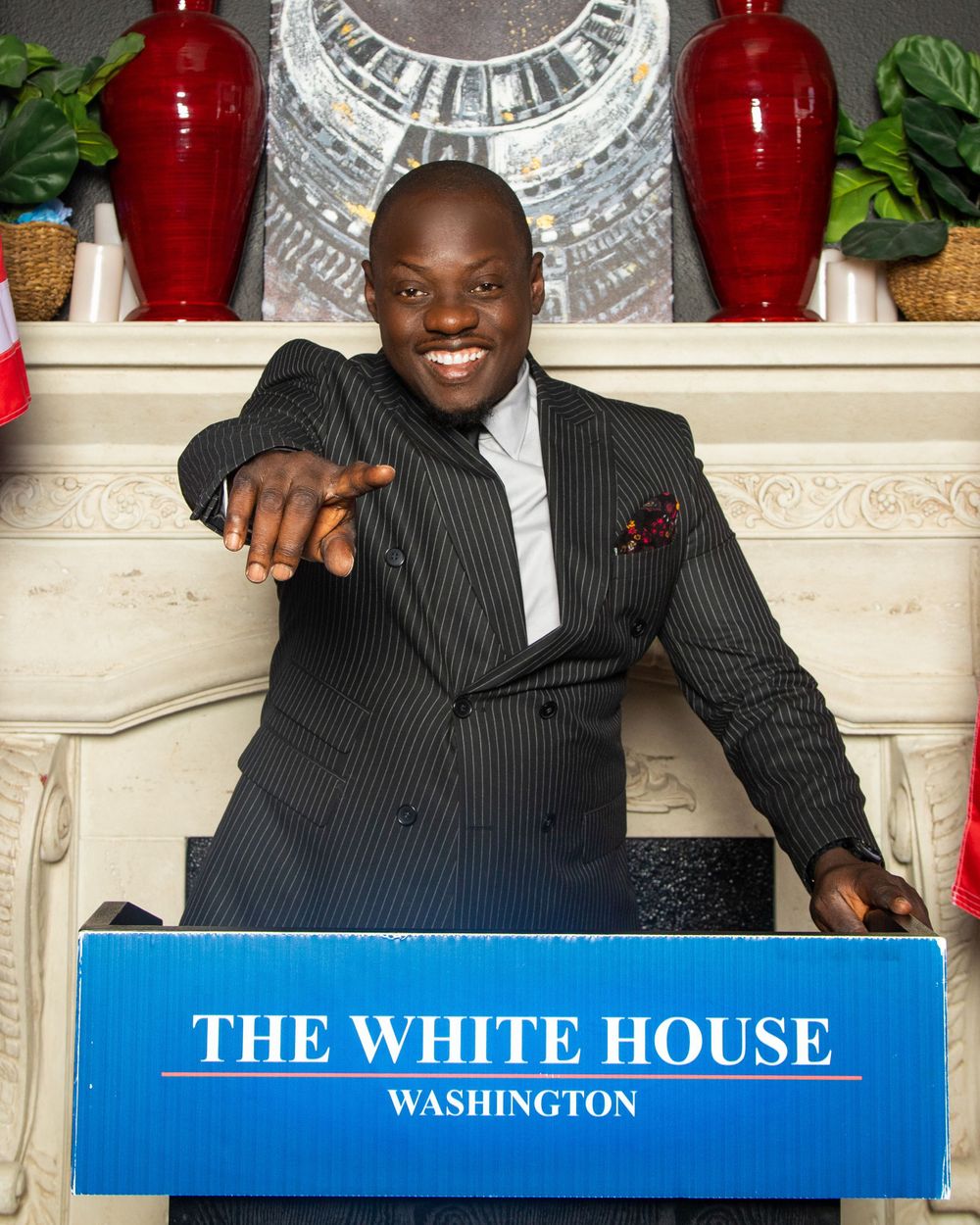 A person in a pinstriped suit poses behind a White House podium with decorative red vases in the background.