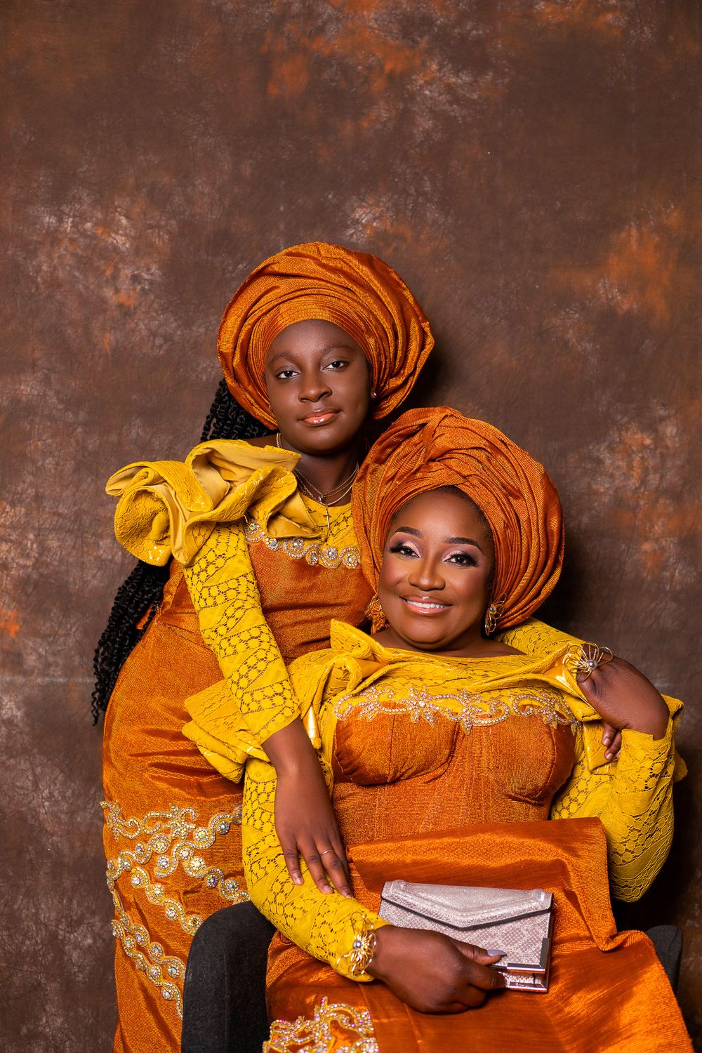 Two people wearing traditional orange and yellow African attire pose against textured brown background.