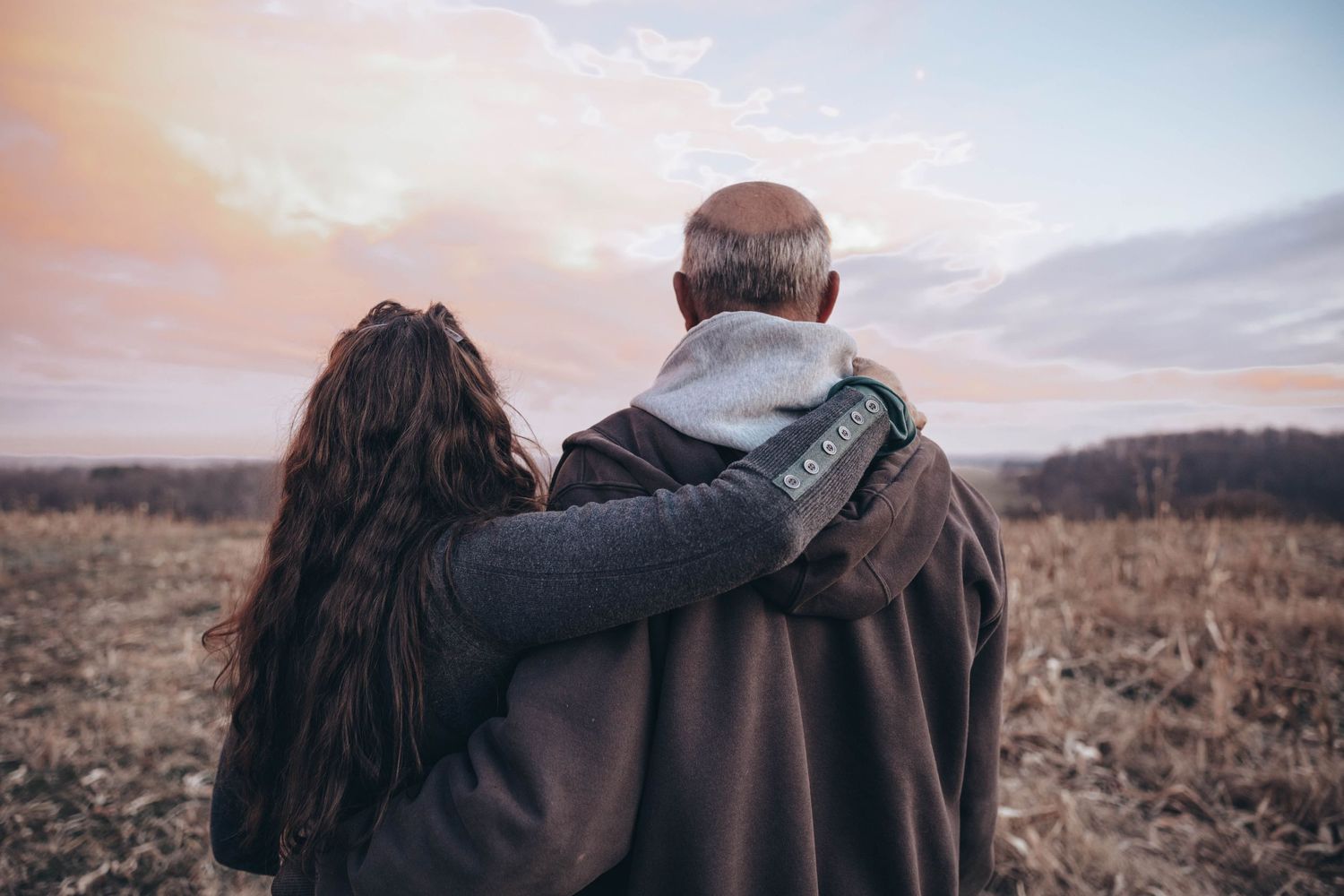 Couple embracing while watching sunset over autumn field from behind.