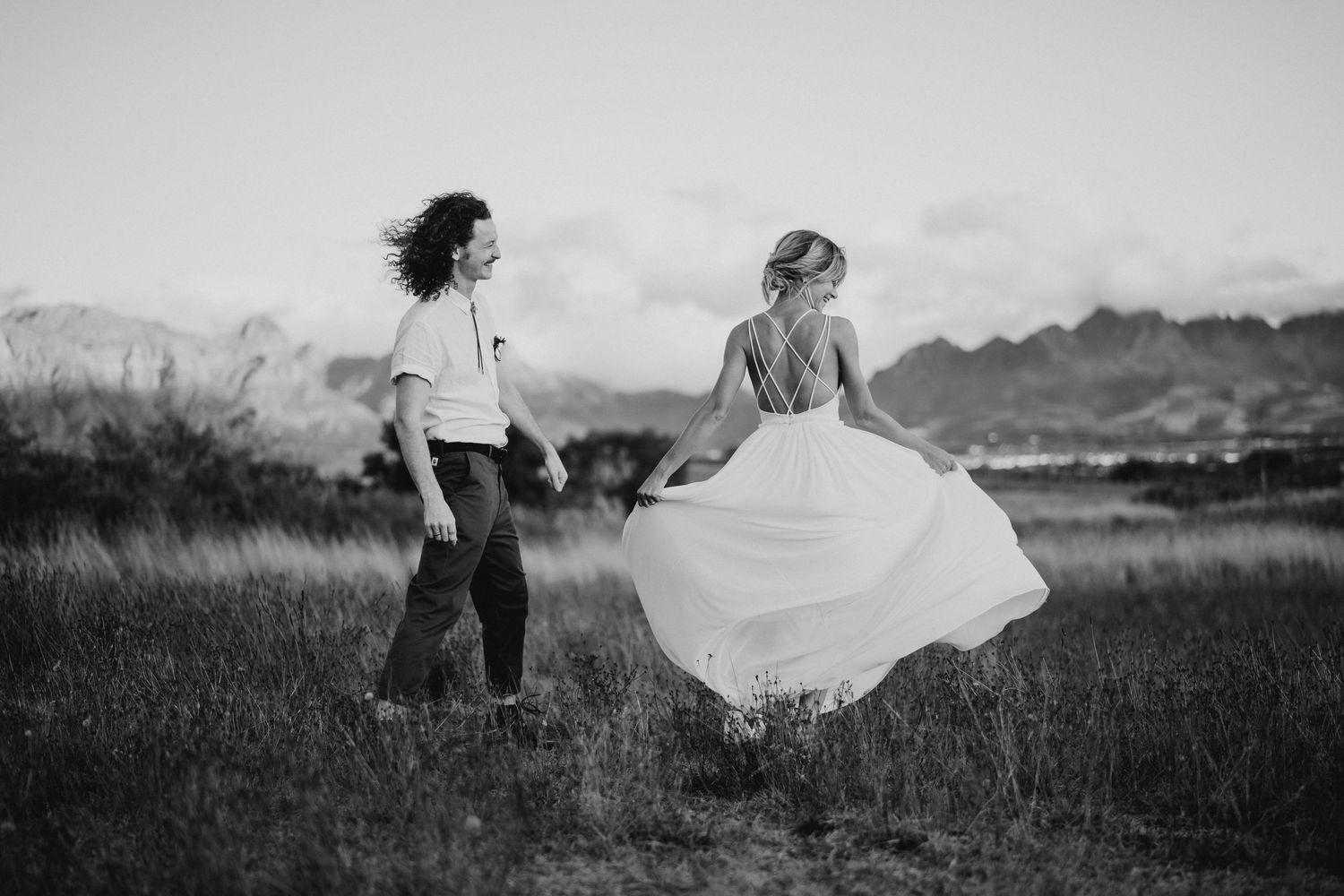 Two people stand in a field with mountains, one wearing a flowing white dress that catches the wind.