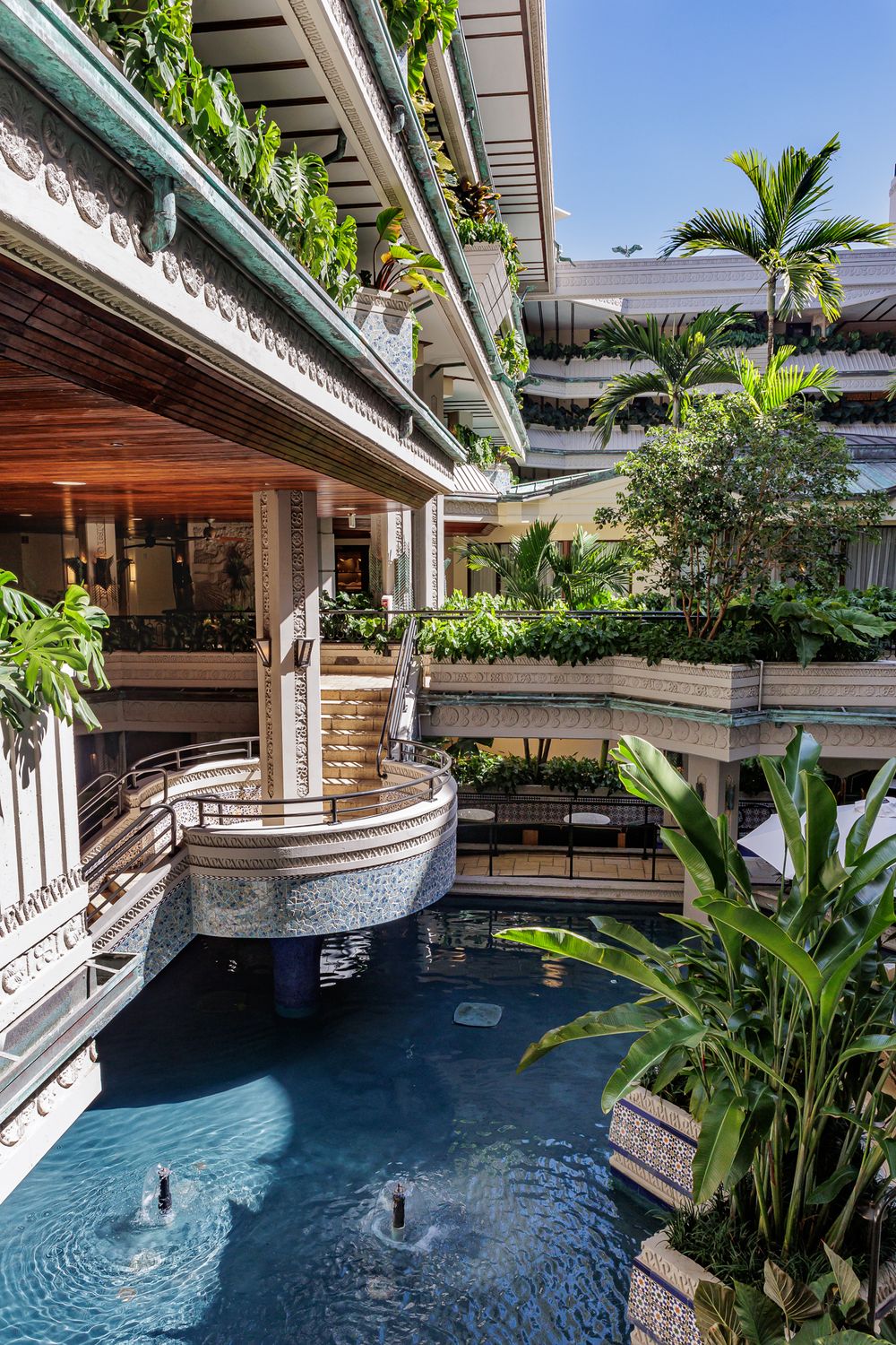 Multi-level open-air atrium with water features and lush tropical plants surrounding a reflecting pool.