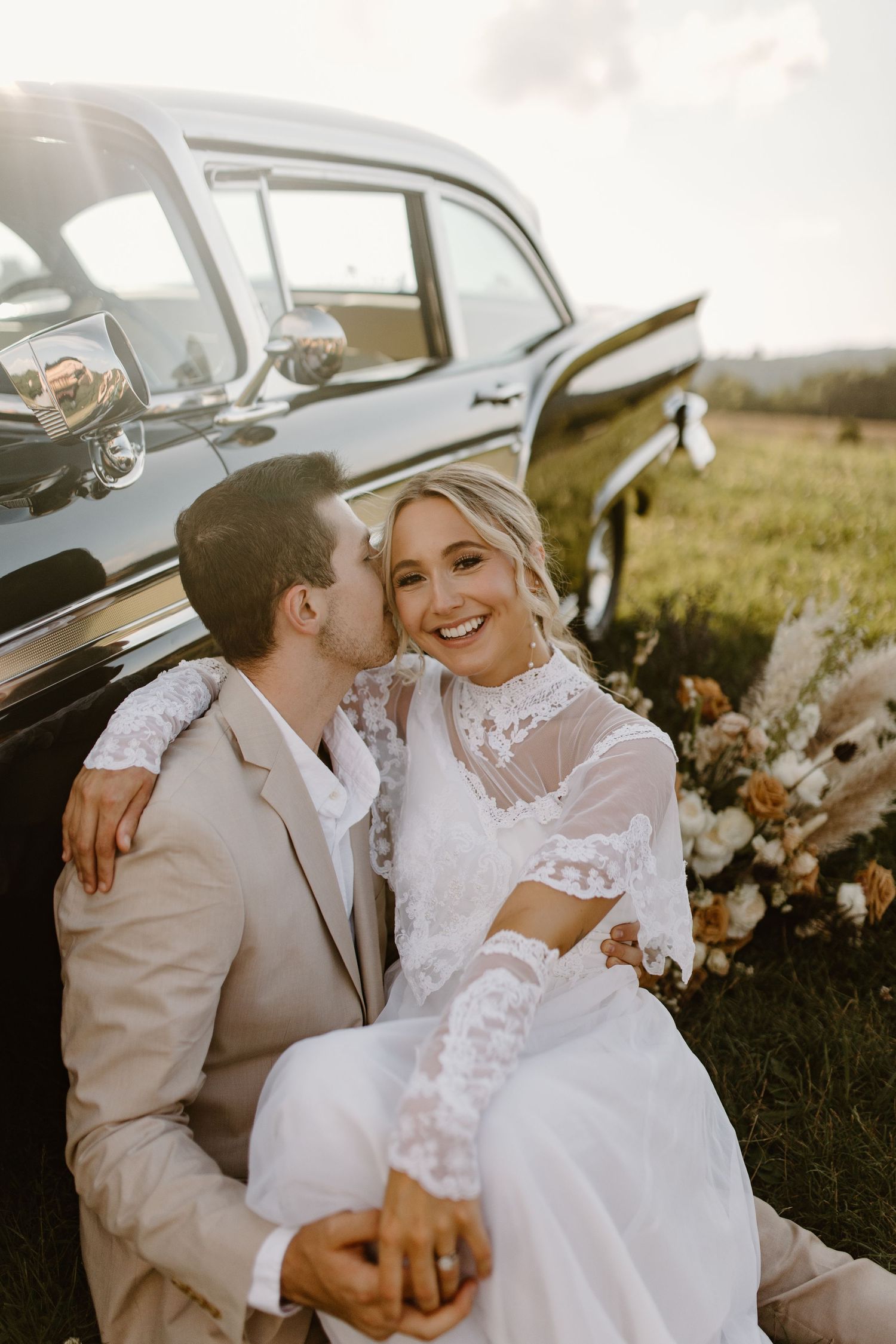Sweet wedding photo captured against a vintage black car in golden afternoon light.