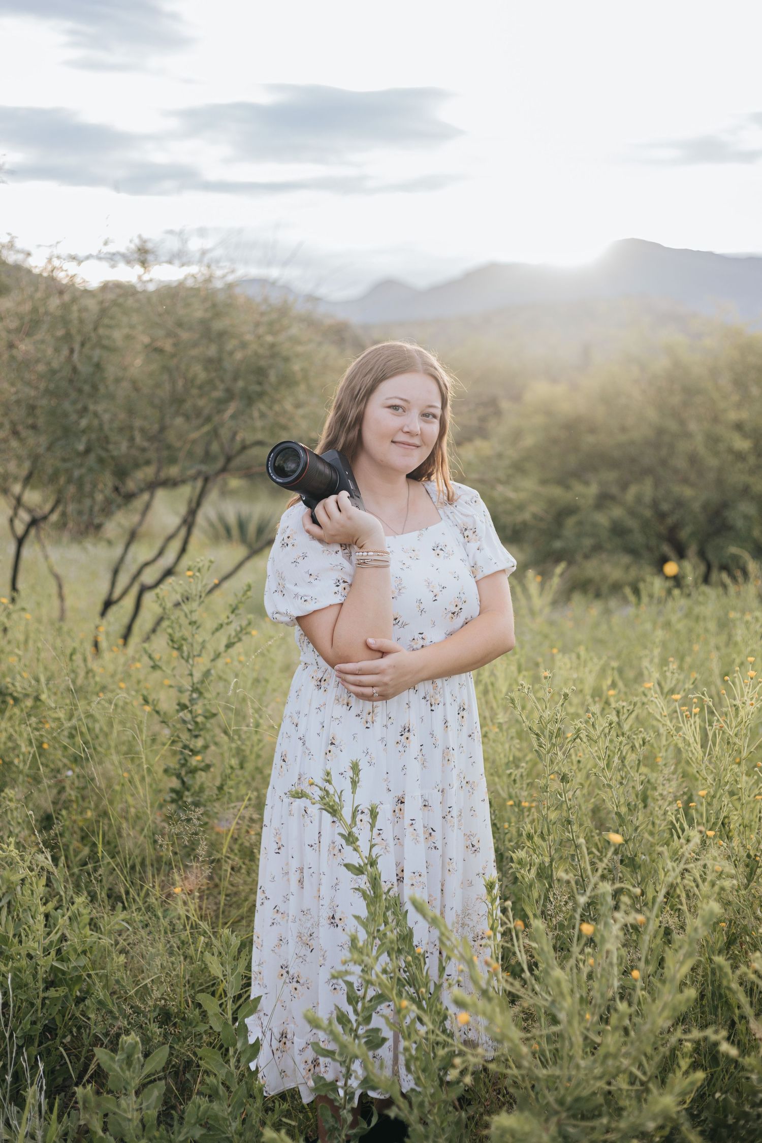 Person in white floral dress standing in wildflower field with mountains in background.