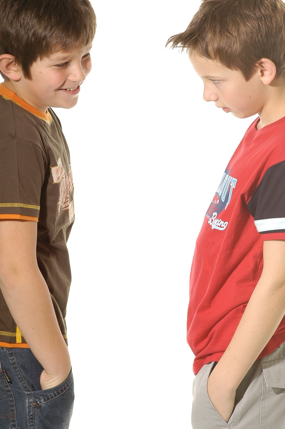 Two young kids in casual clothes standing face to face with hands in pockets against white background.