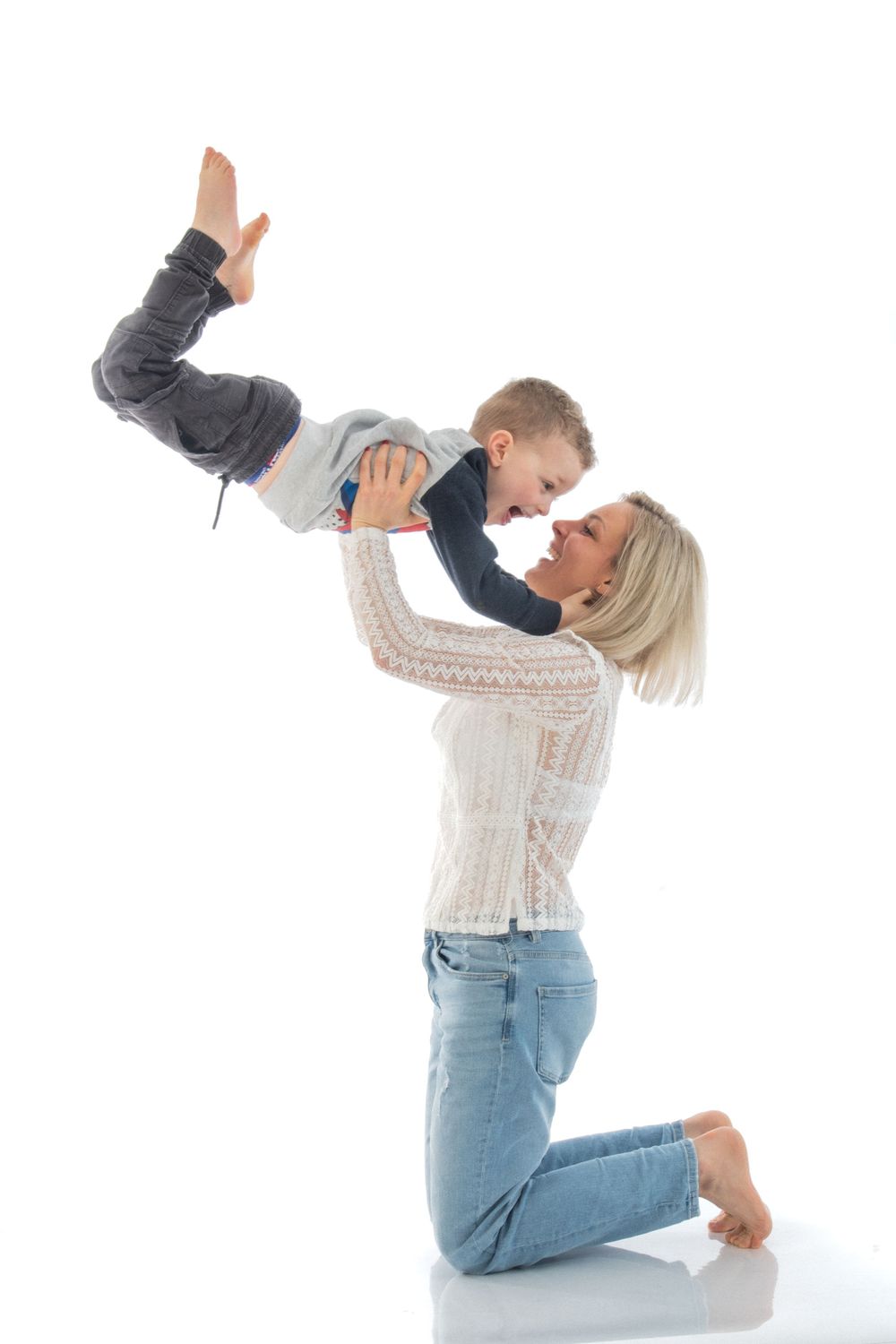 A parent lifts a small child playfully in the air while kneeling on a white background.