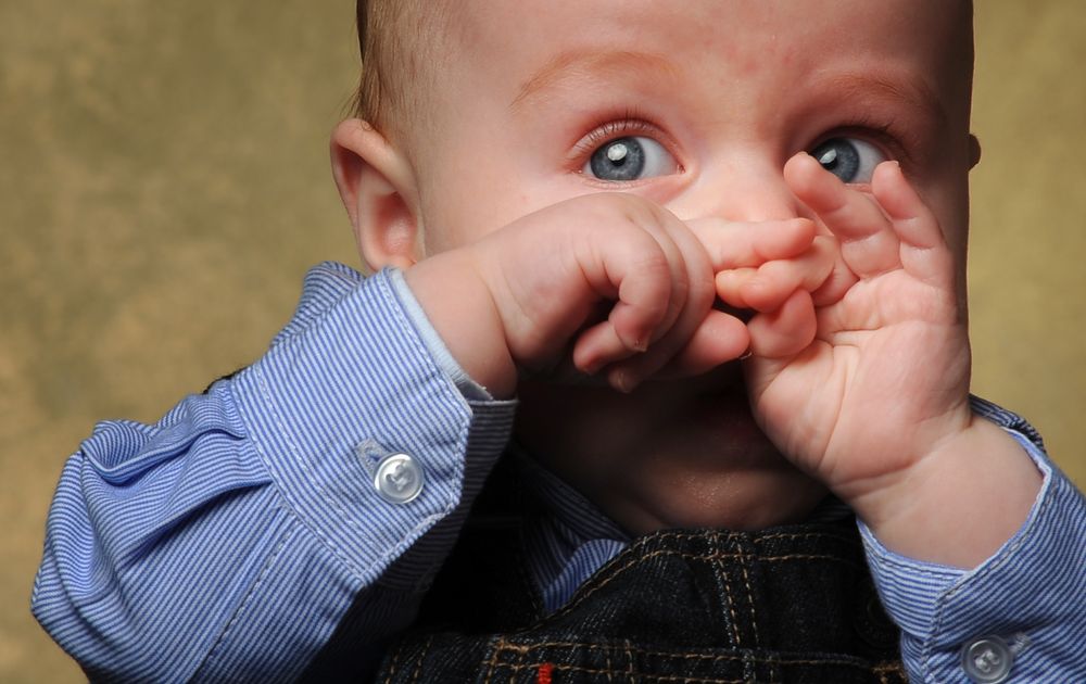 Close-up of baby in blue shirt rubbing eyes with tiny hands.