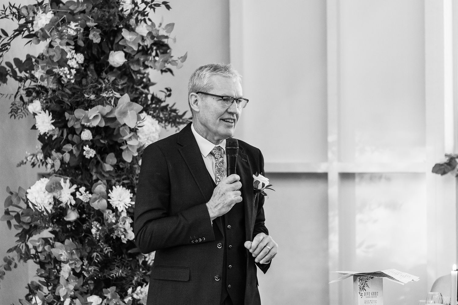 Black and white photo of speaker delivering wedding speech beside floral arrangements.