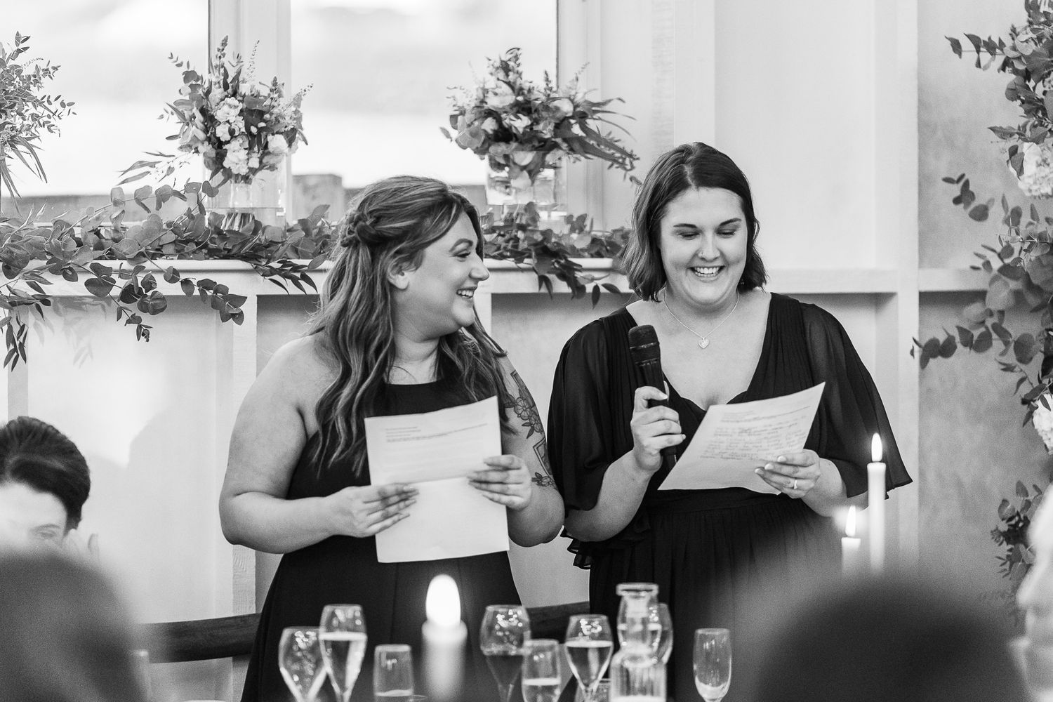 Black and white photo of wedding guests delivering speeches at reception table.
