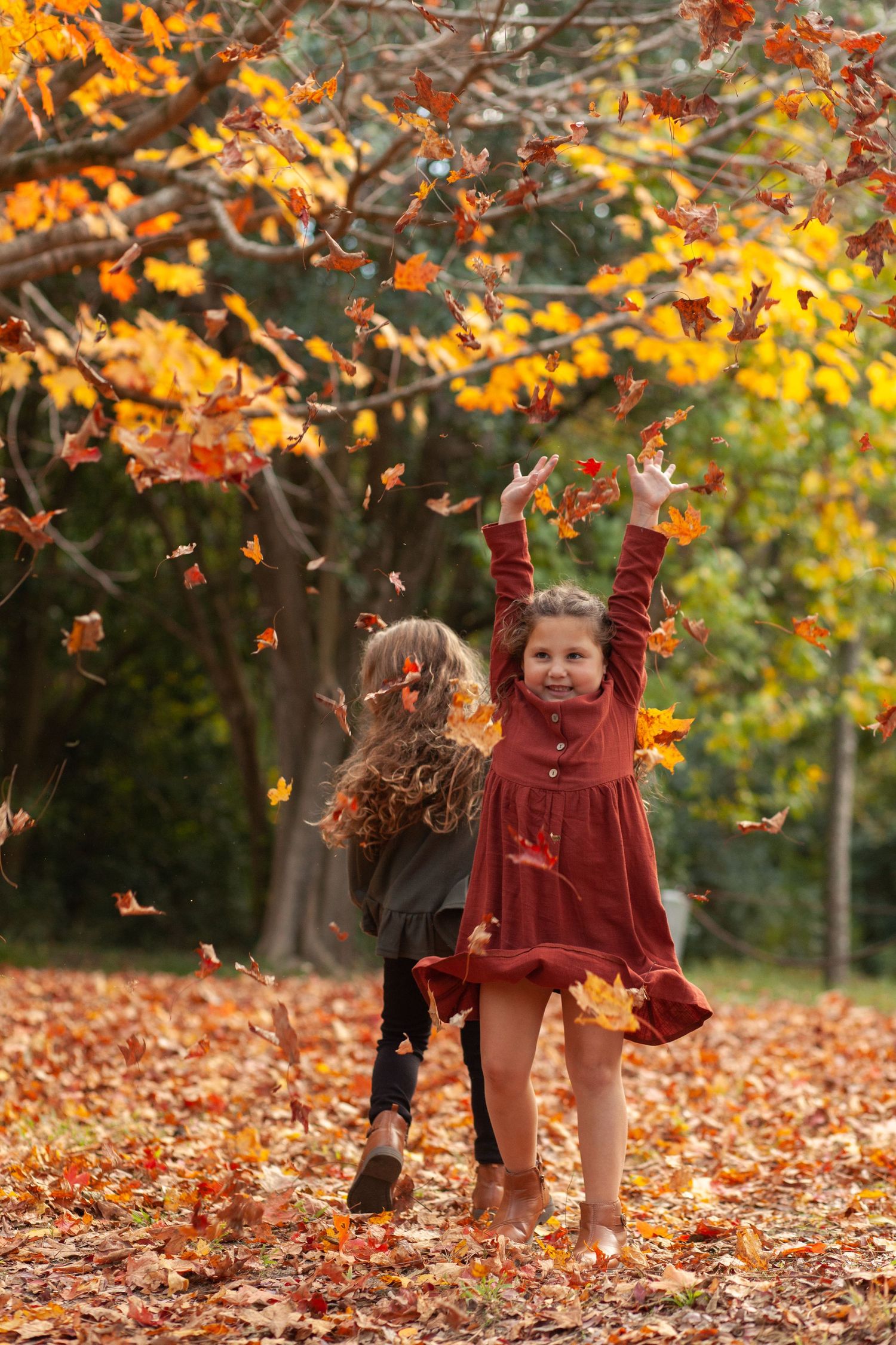 Two children play in fallen autumn leaves under trees with vibrant orange and yellow foliage on a fall day.