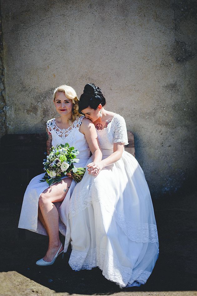 Two brides in white dresses sitting together against a sunlit wall with wedding bouquet.