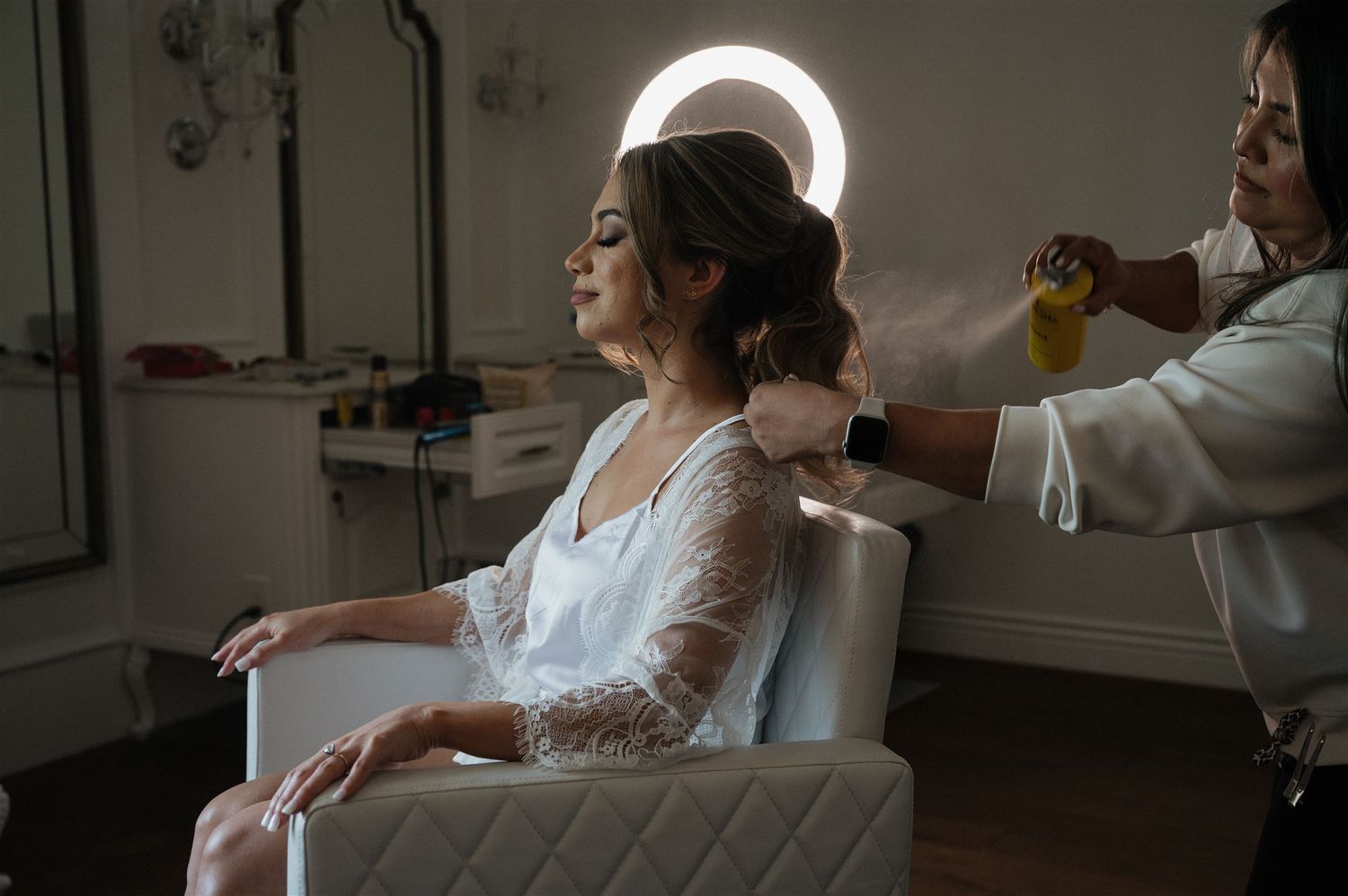 Someone sits in a chair getting hair styled at a salon with a glowing circular light fixture visible in the background.