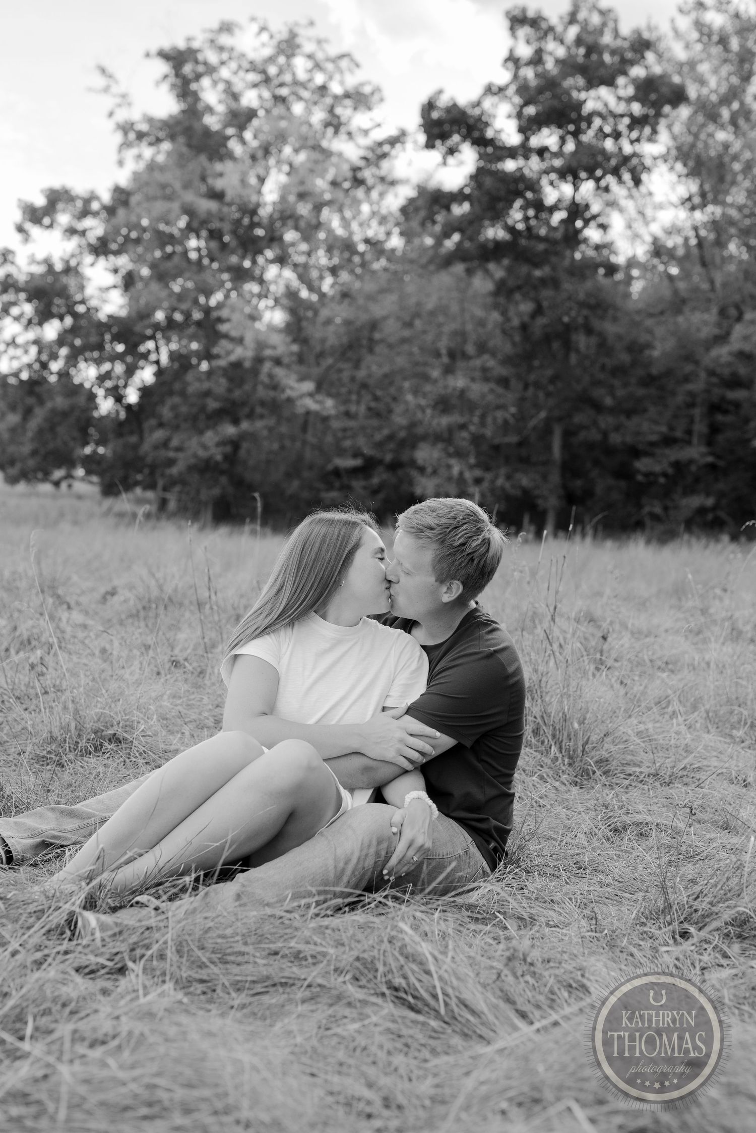 Intimate black and white portrait of a couple cuddling together in a grassy meadow with trees in background.