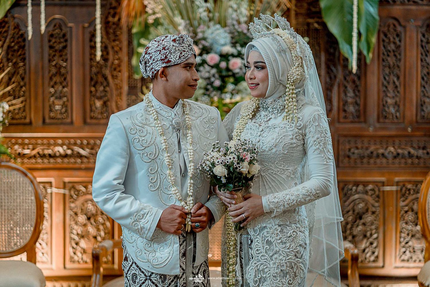 Traditional Javanese wedding ceremony with ornate wooden backdrop and couple in matching white attire.