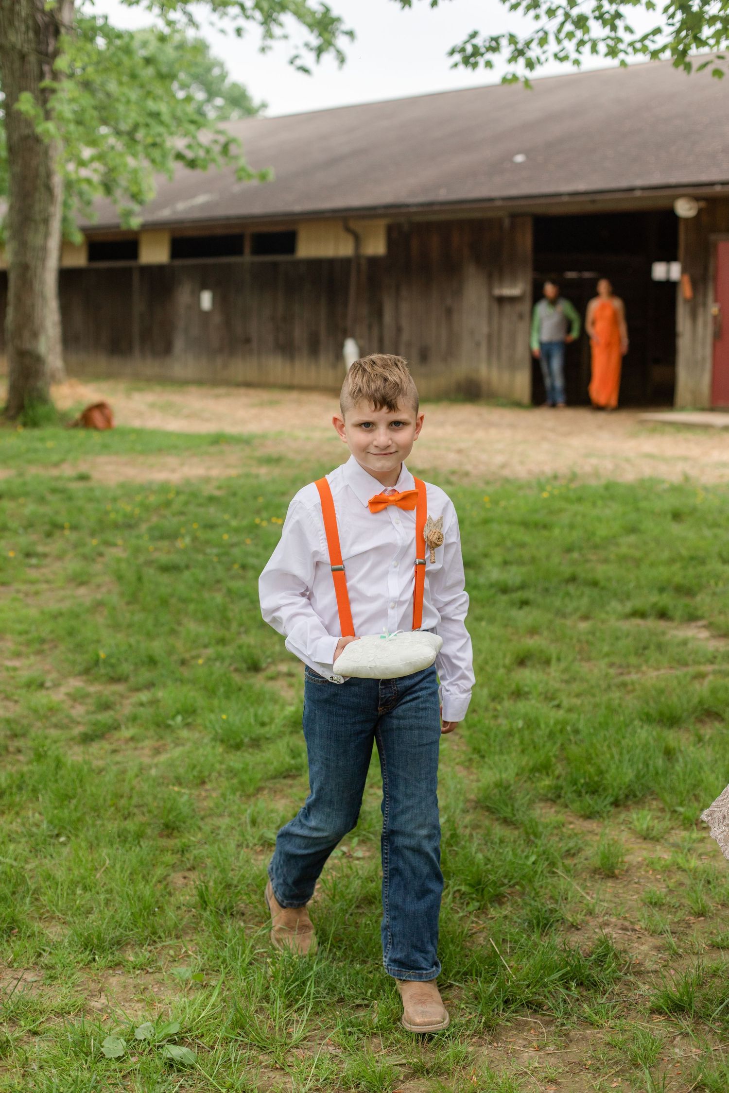A young attendant in suspenders and bow tie stands on grass near a wooden building.